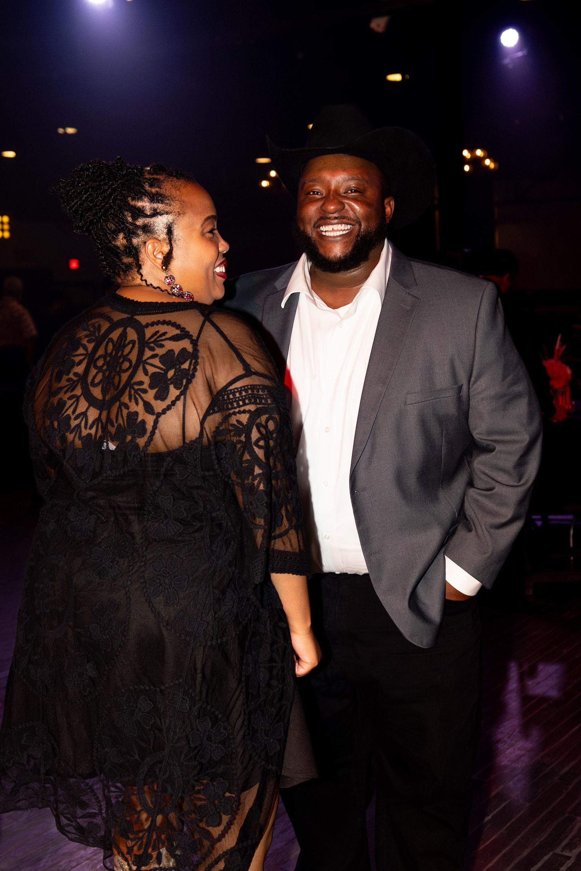 Woman in black lace dress, man in gray blazer smile at an event.