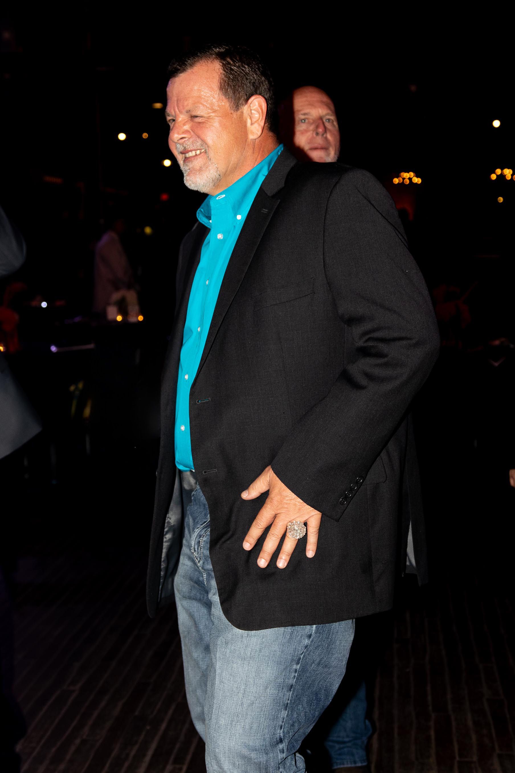 Man in a blazer and jeans, smiling, indoors. A second person is visible behind him.