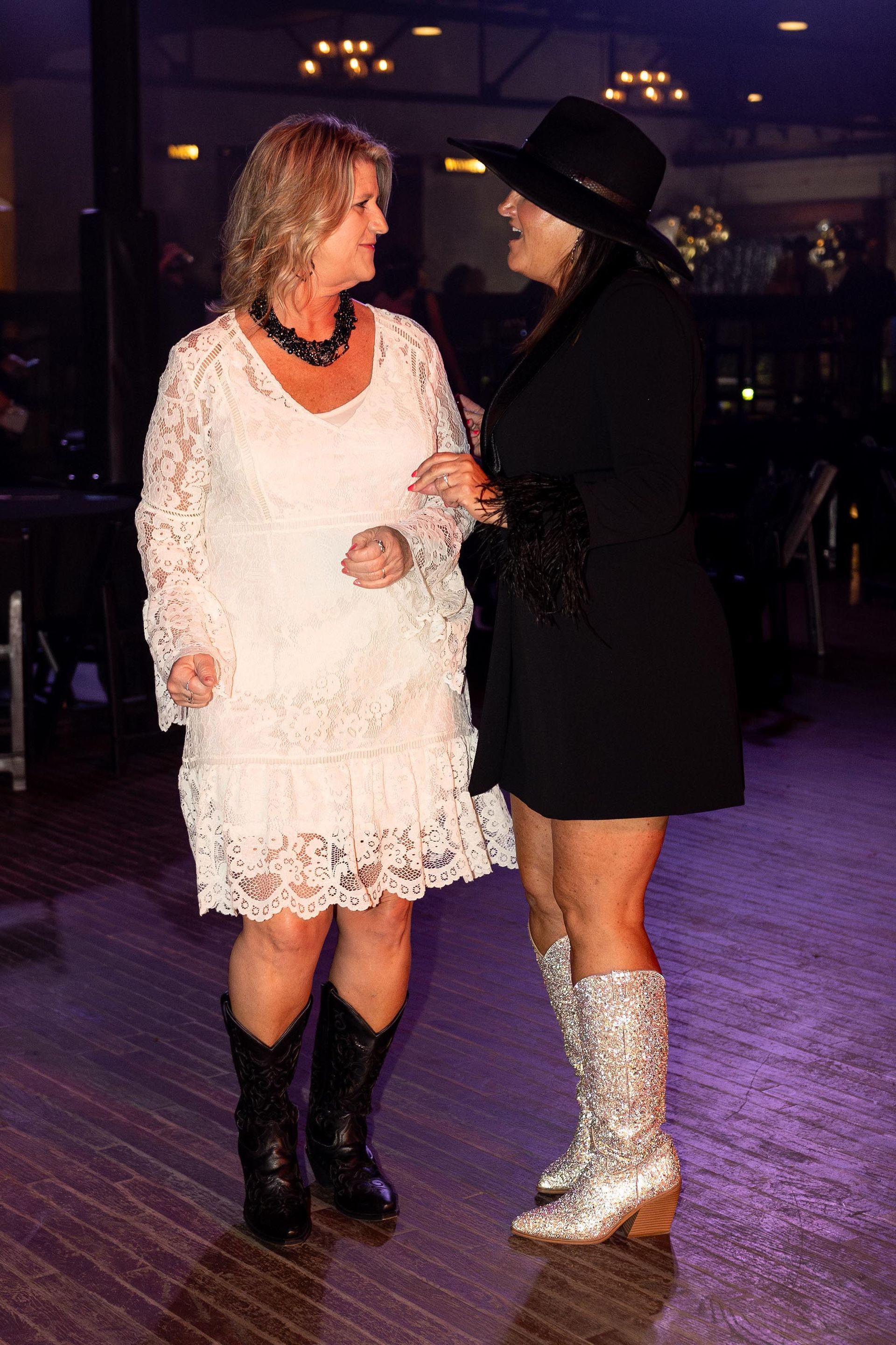 Two women in a dim setting, conversing. One in white dress, black boots. Other in black outfit, cowboy hat, silver boots.