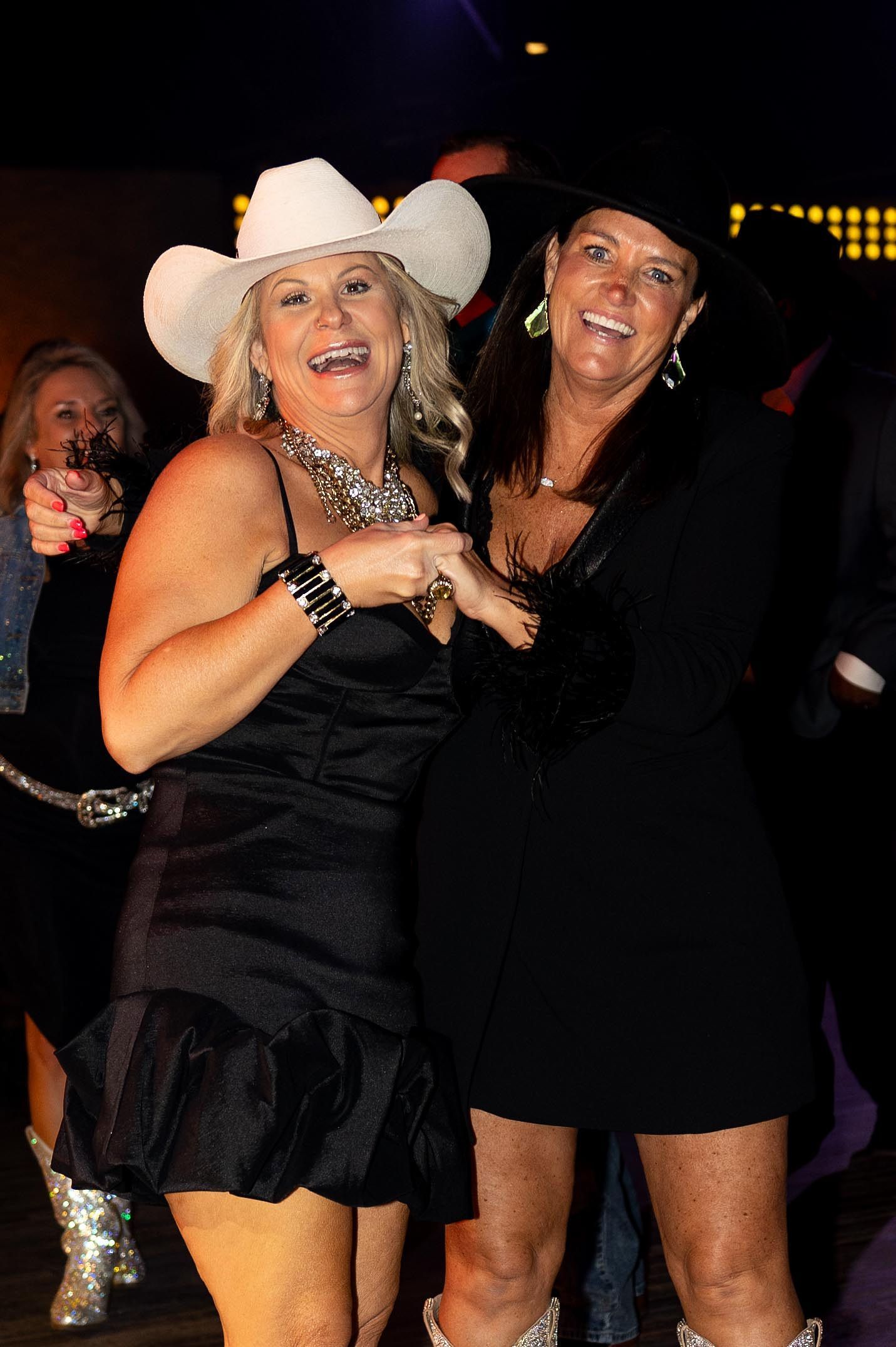 Two women in cowboy hats laugh, one in a black dress, the other in a white dress.