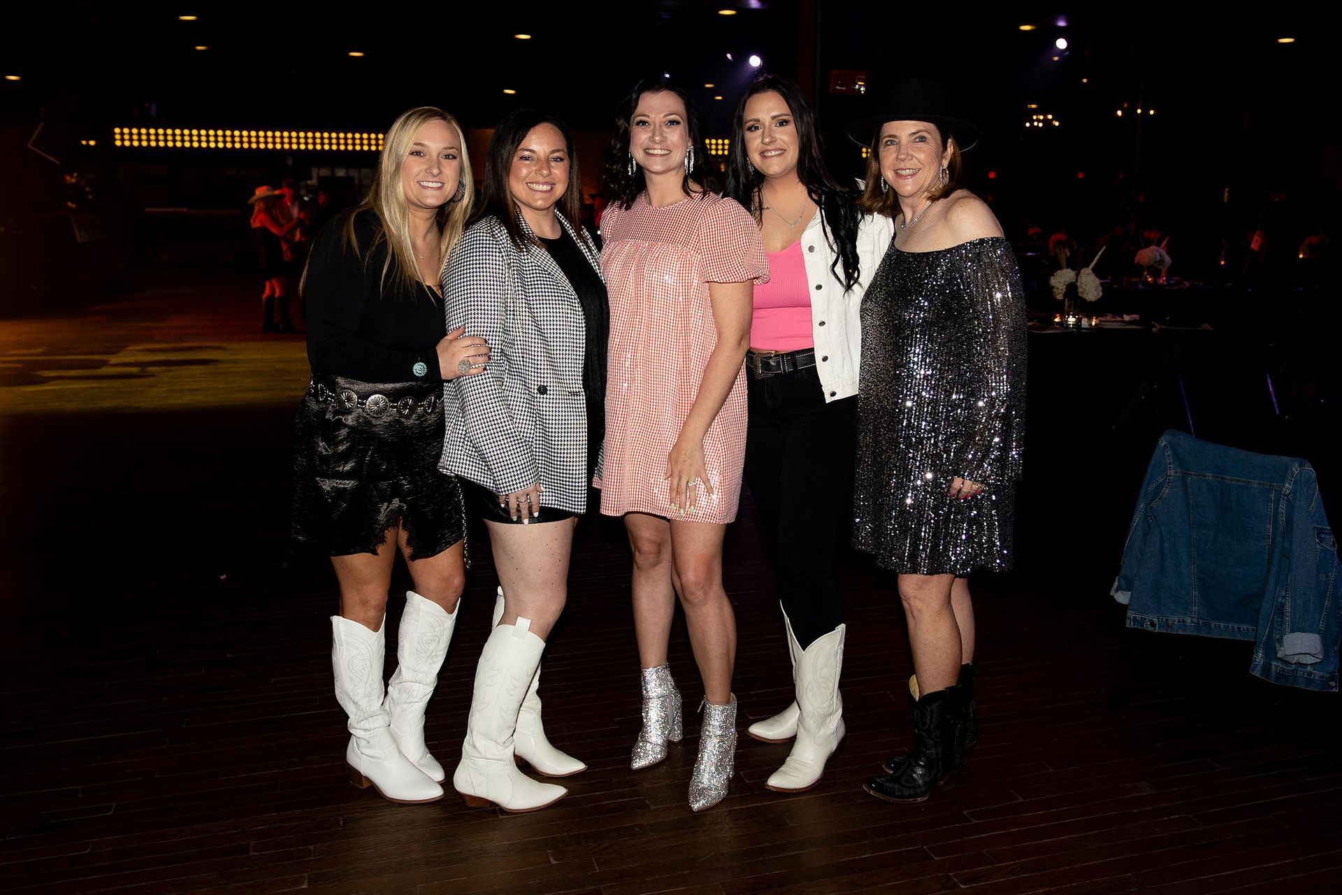Five women in boots pose indoors. One wears an off-the-shoulder sequin dress, others wear dresses and jackets.
