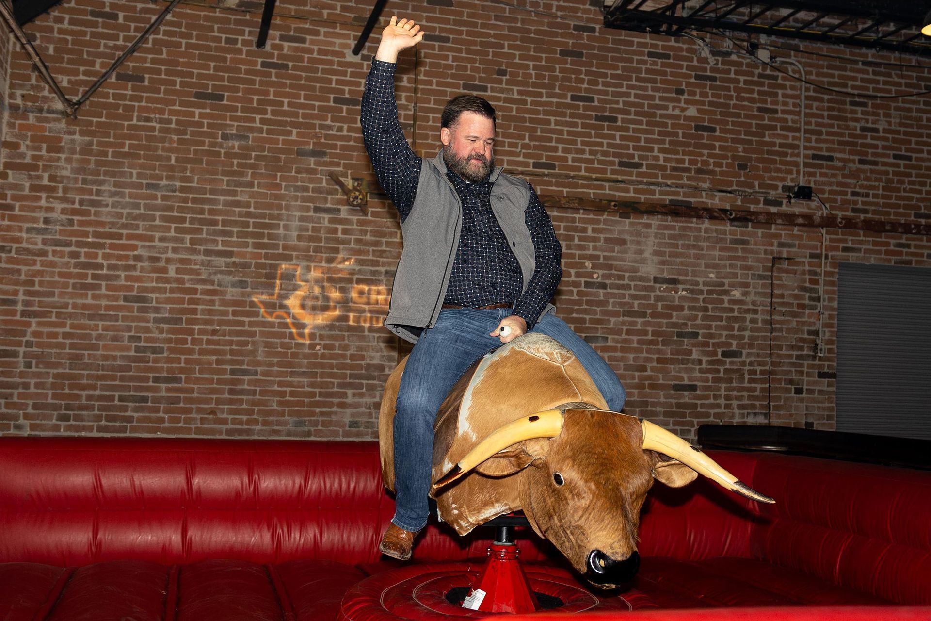 Man on a mechanical bull, raising arm, brick wall backdrop.