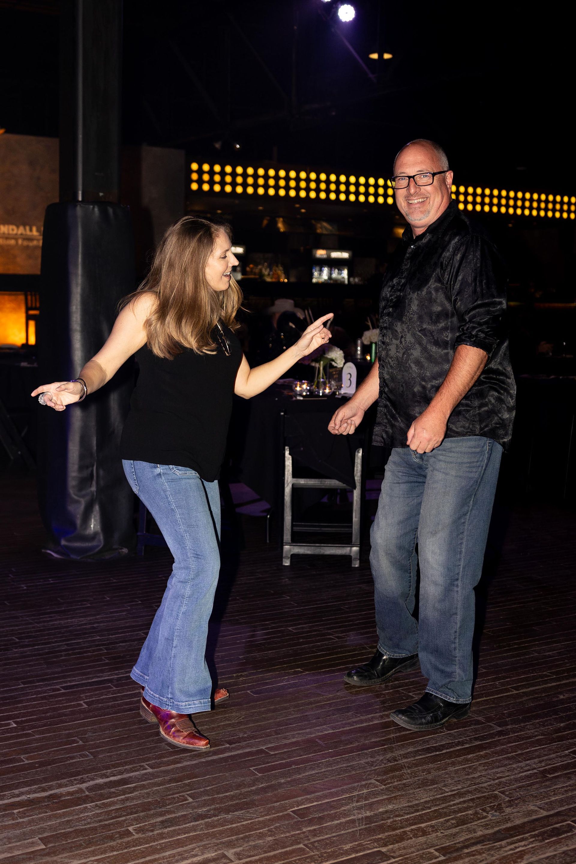 A woman and a man dancing at a dimly lit event; woman in jeans and black top, man in jeans and black shirt.