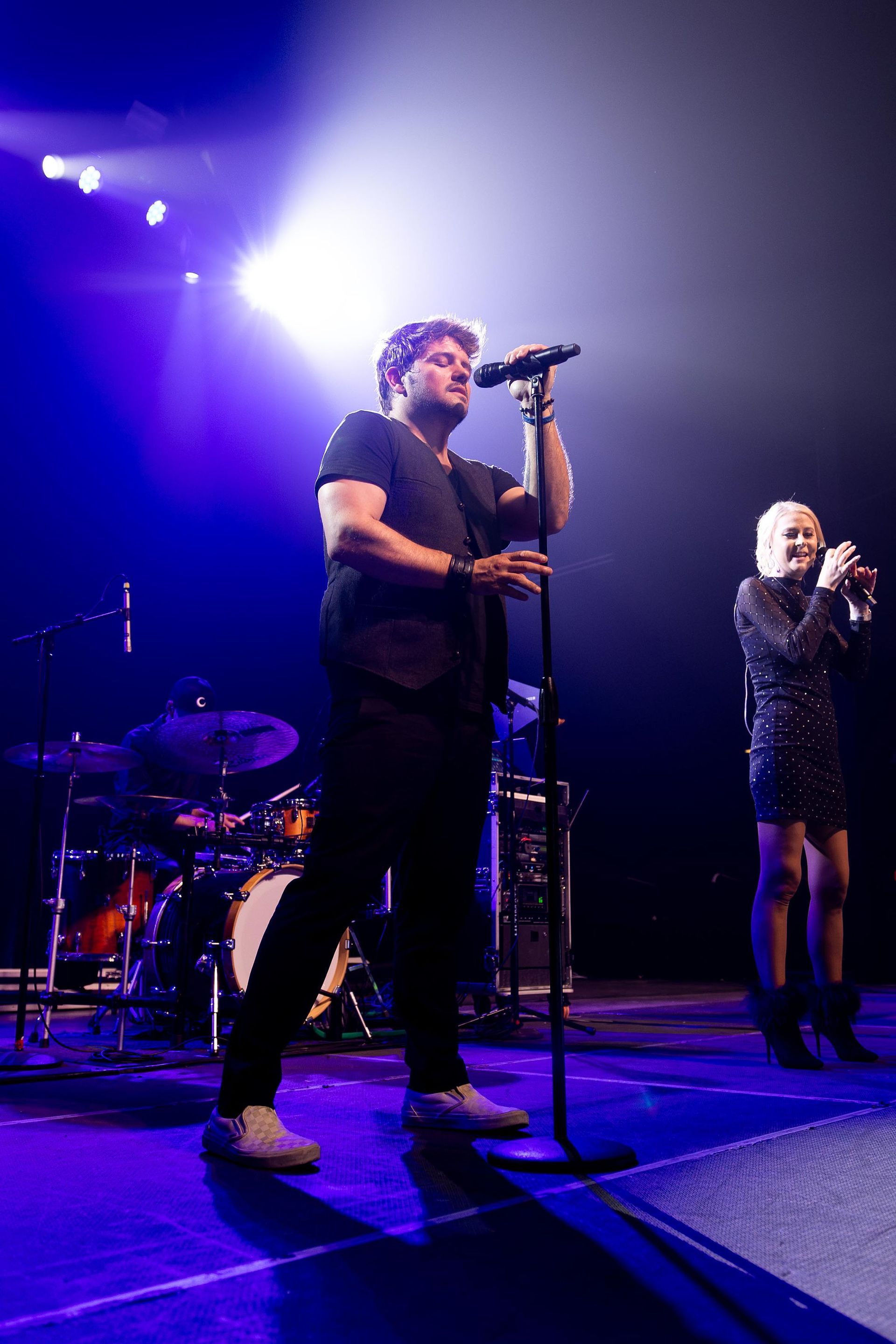 Two performers on stage, man singing into a mic, woman in sequin dress; concert setting, blue light.