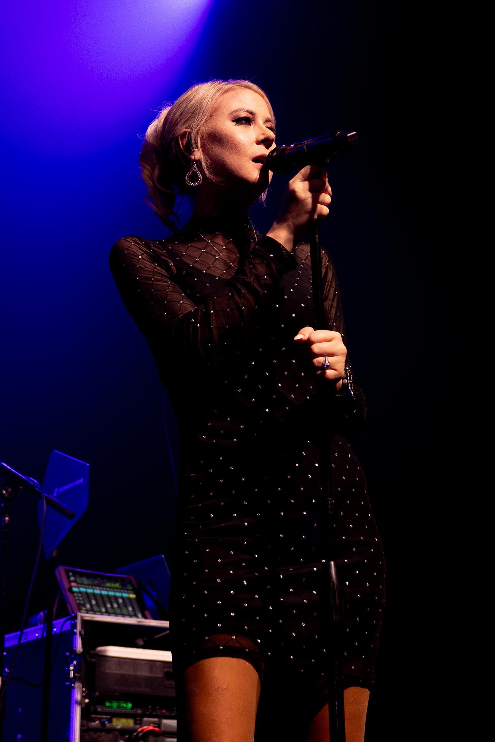 Blonde woman singing on stage, wearing a black dress with sparkling details.