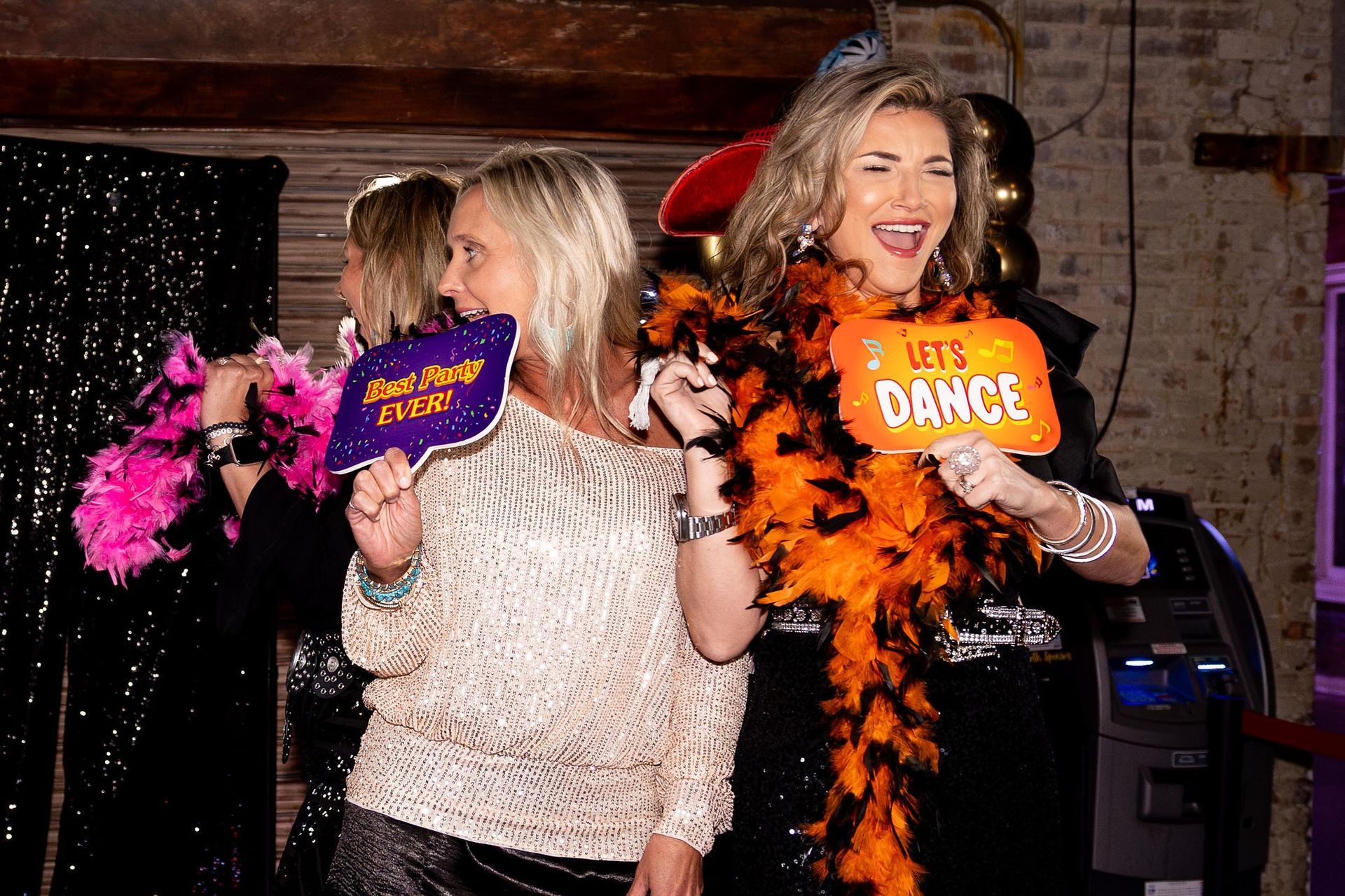 Two women at a party, smiling, holding photo booth props, and wearing boas.