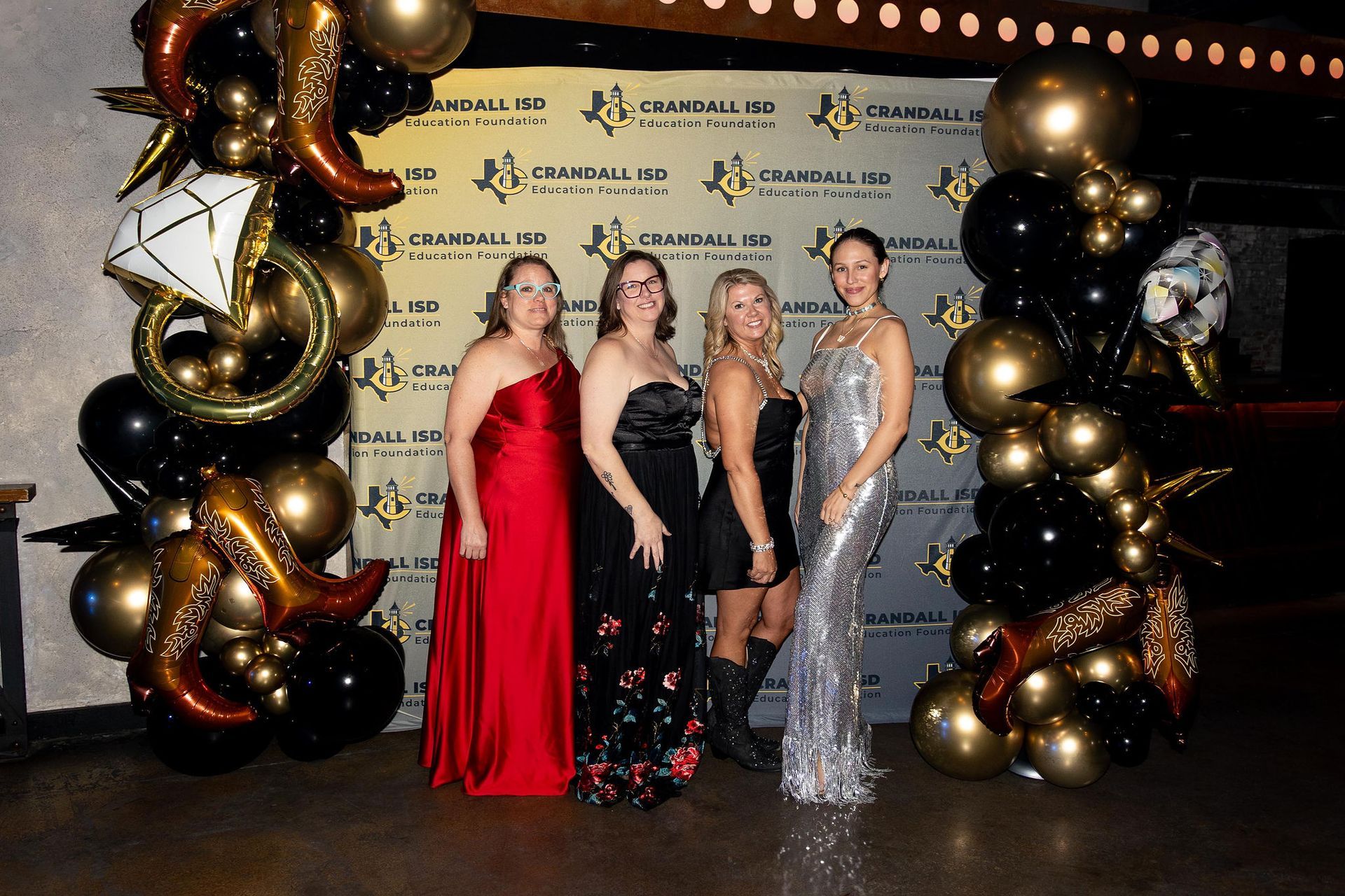 Four women in formal wear pose at a gala event with balloon archway; black and gold balloons, western boots.