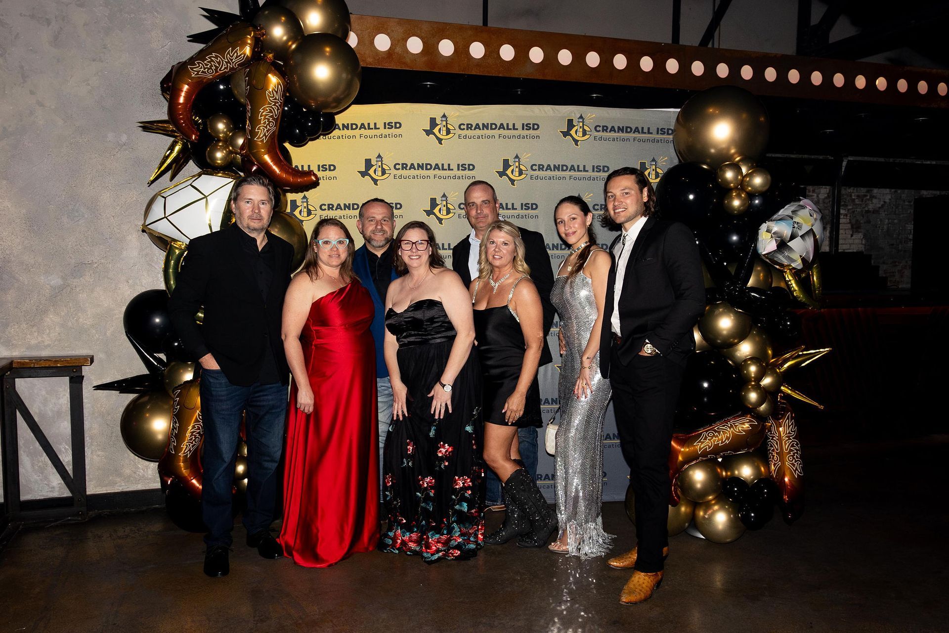 Group of people posing at an event, with balloons and a backdrop. Formal attire, smiling.