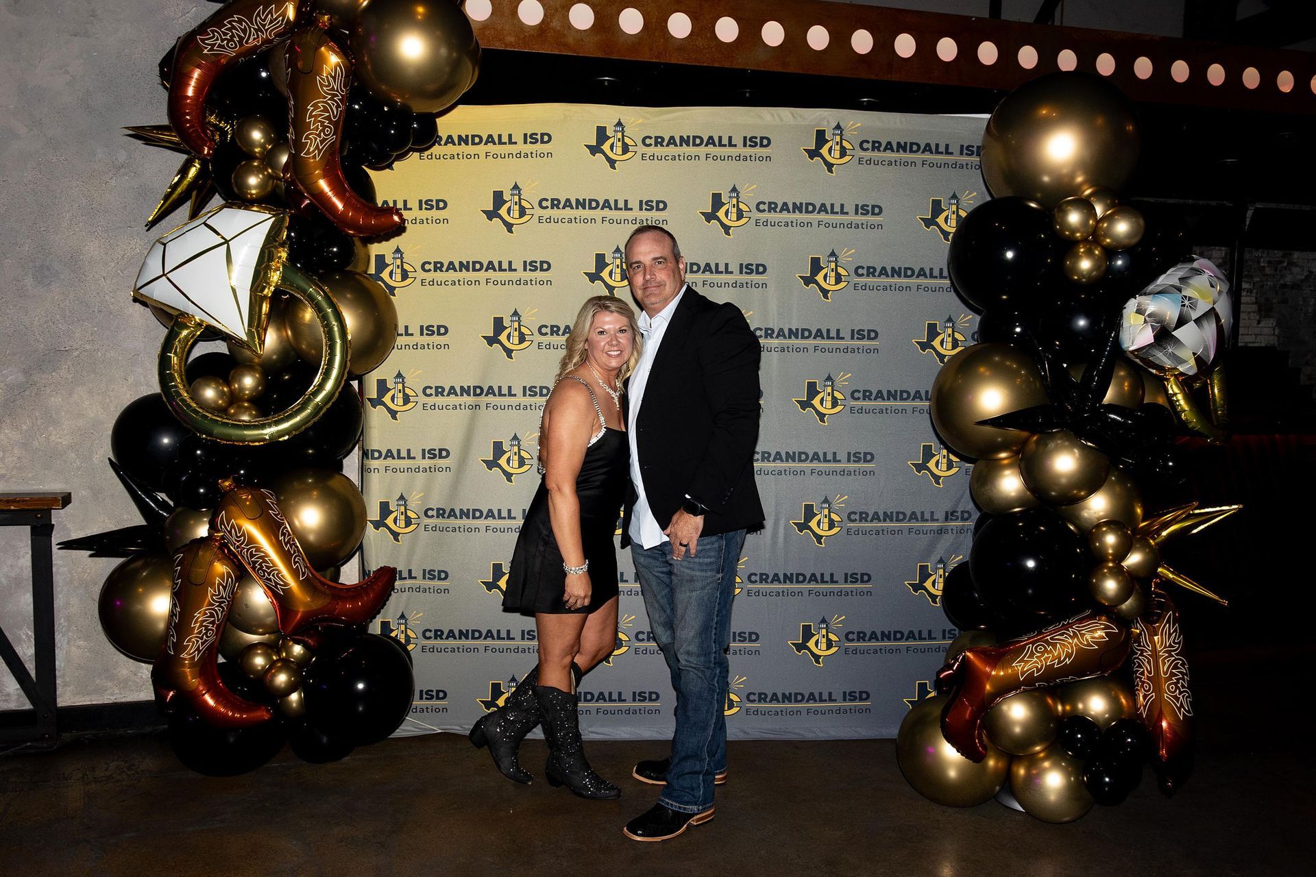 Couple posing in front of a backdrop with gold and black balloons at an event.