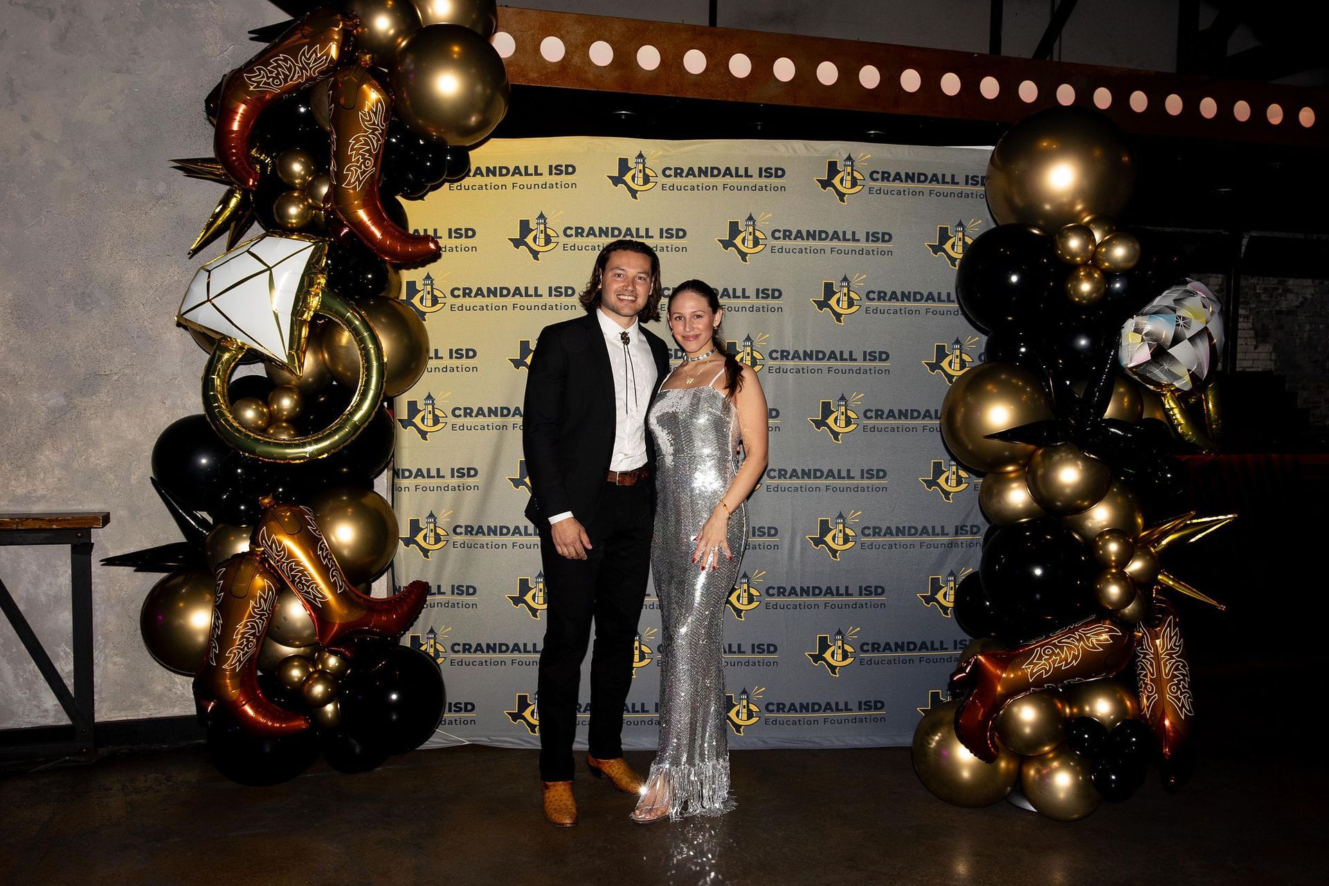 Man and woman pose in front of a branded backdrop, surrounded by balloons. They are dressed formally.