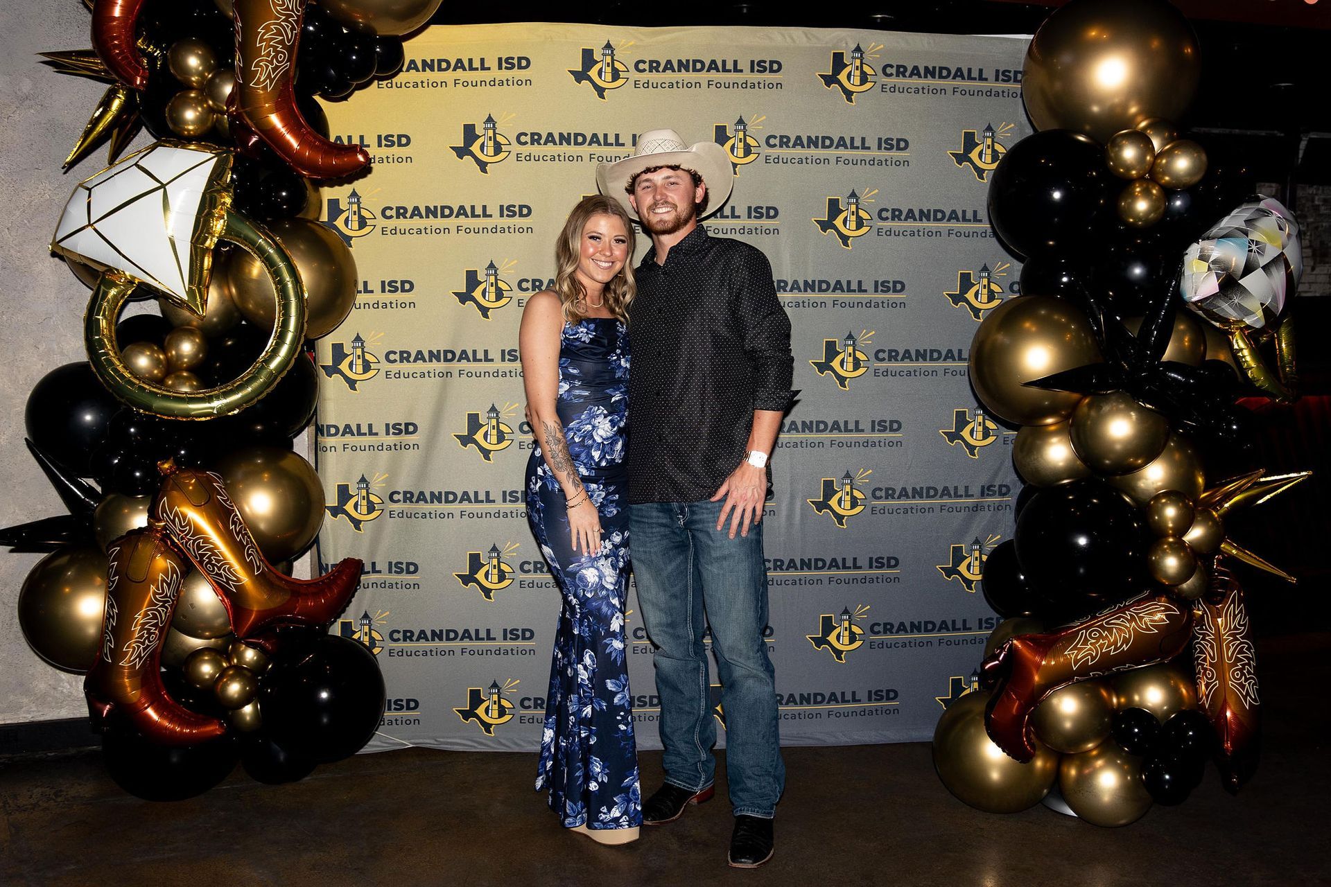 Couple posing in front of a branded backdrop with balloon decorations; woman in blue gown, man in cowboy hat.