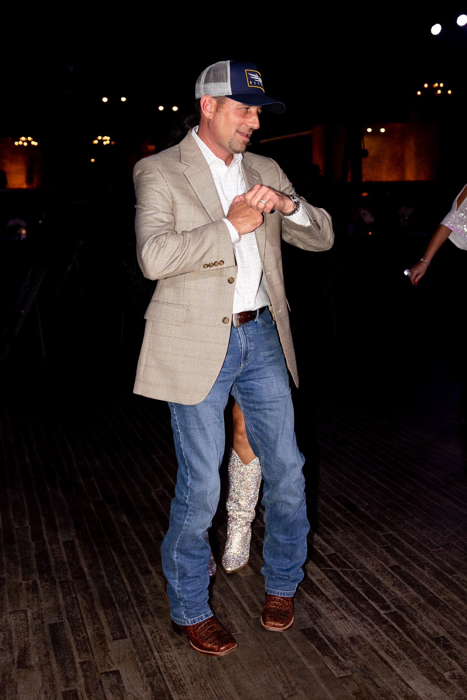 Man in a blazer, jeans, and a baseball cap dancing on a wooden floor.