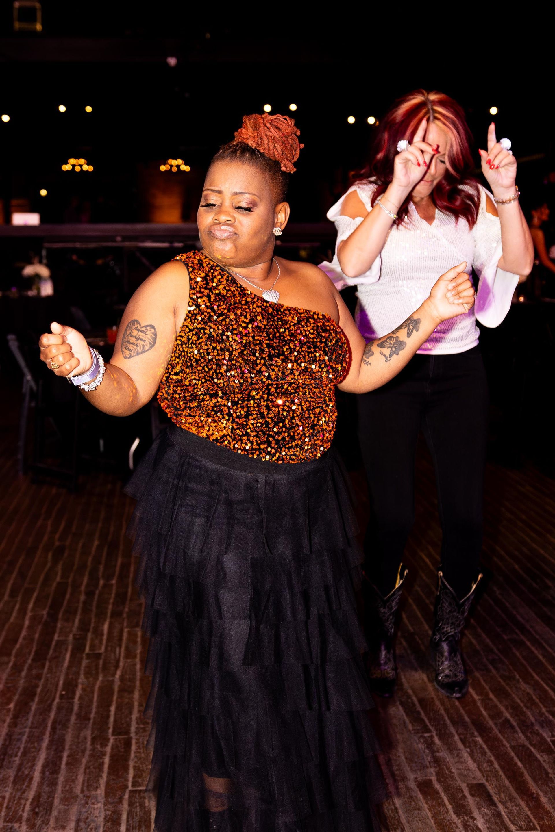 Two women dancing at a dark event. One in a sequin top, black skirt; the other in a white top, black pants.