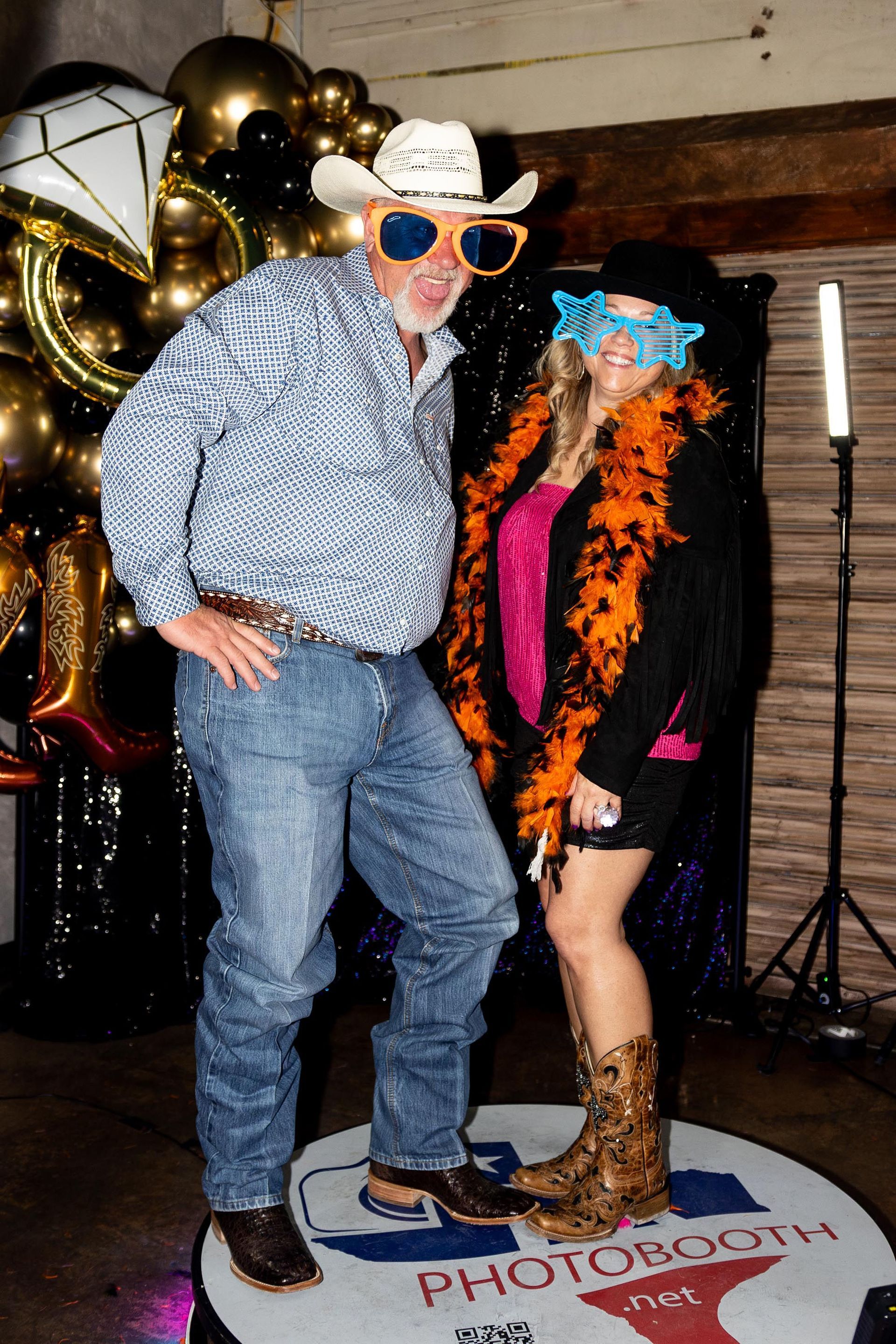 Man and woman posing in a photo booth. Man in cowboy hat and oversized glasses. Woman in orange boa and cowboy boots.