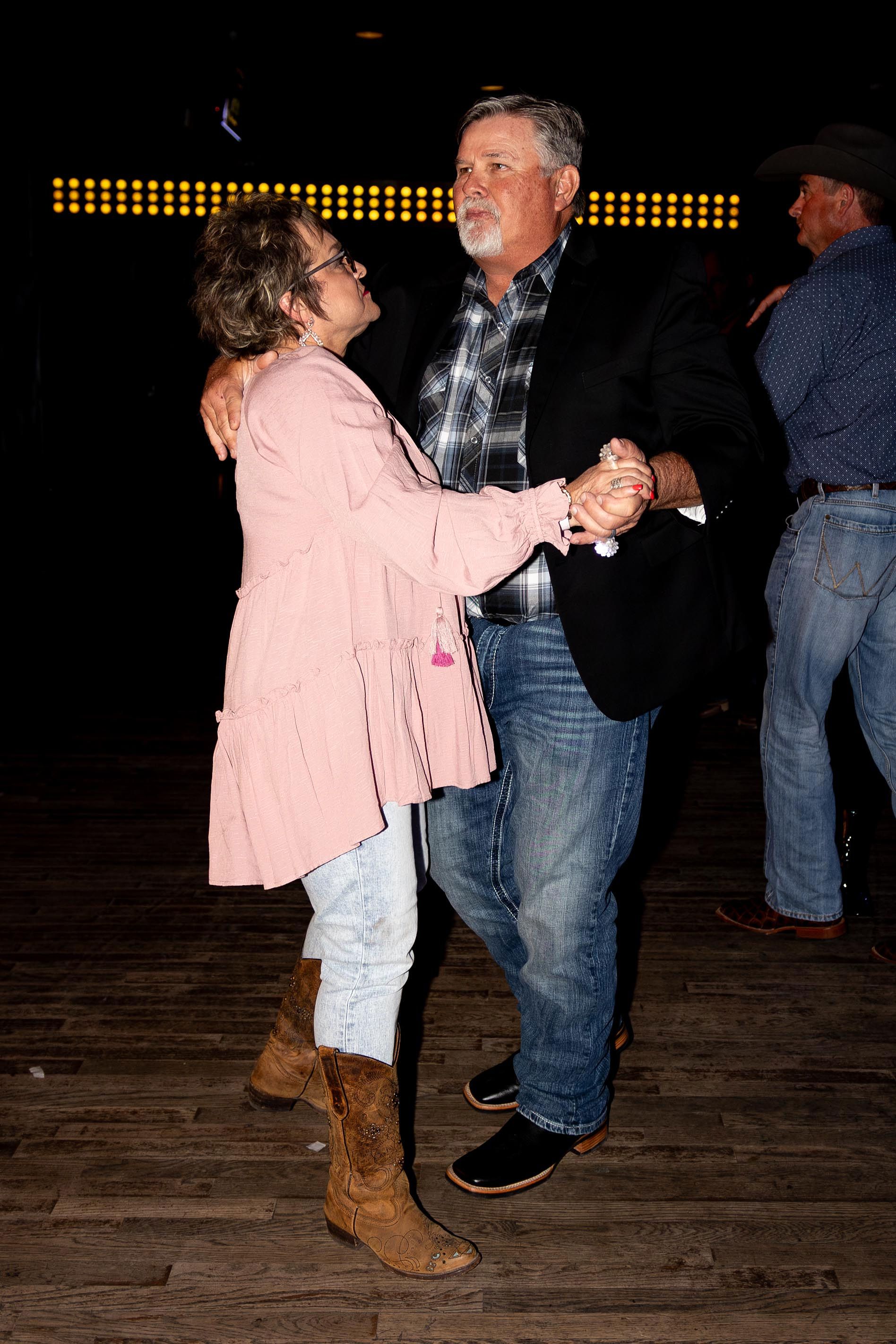 Couple dancing at a venue; woman in pink jacket & boots, man in plaid & jeans; setting is dark with lights.