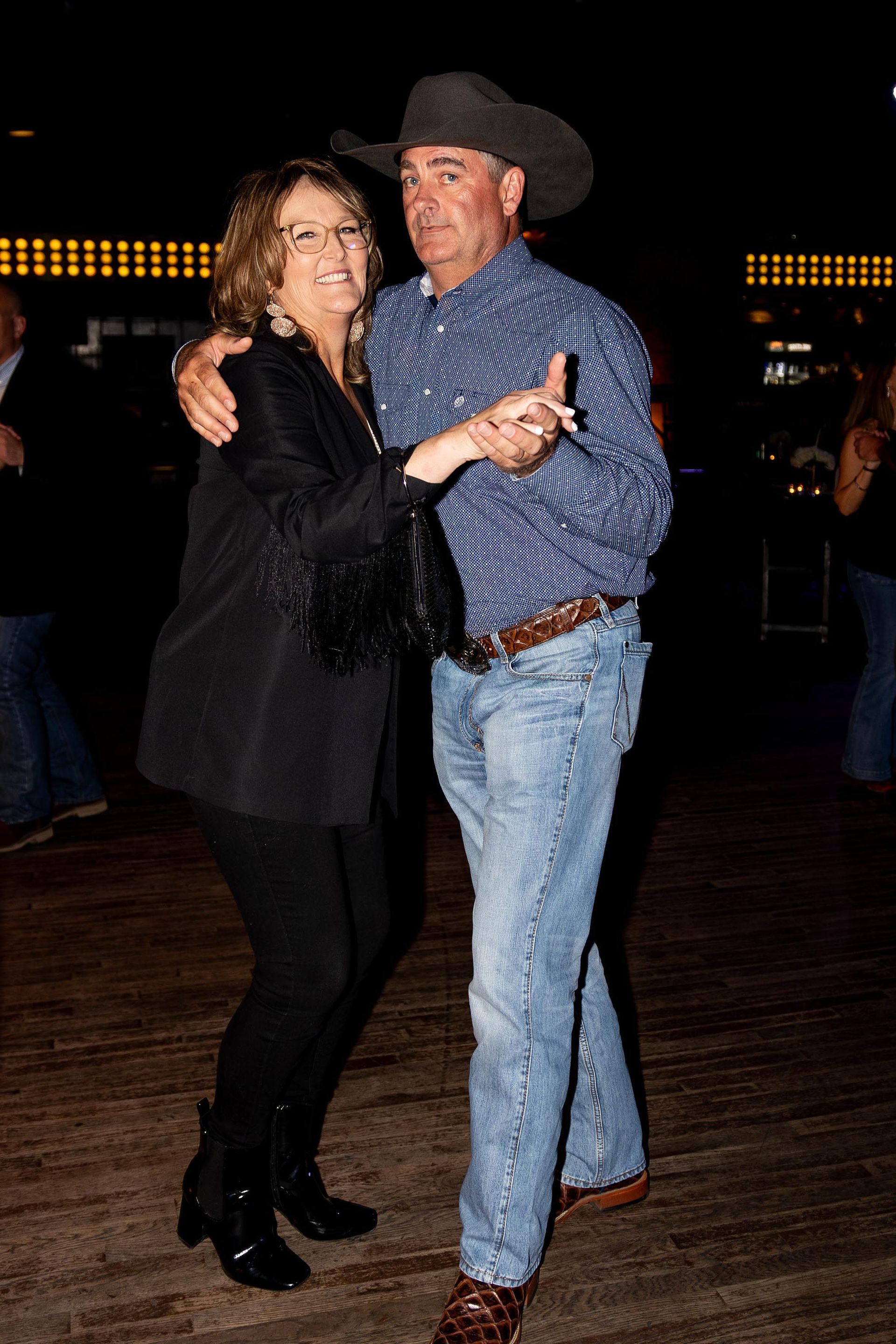 Couple dances in a dimly lit room; the man wears a cowboy hat and shirt, the woman a black jacket.