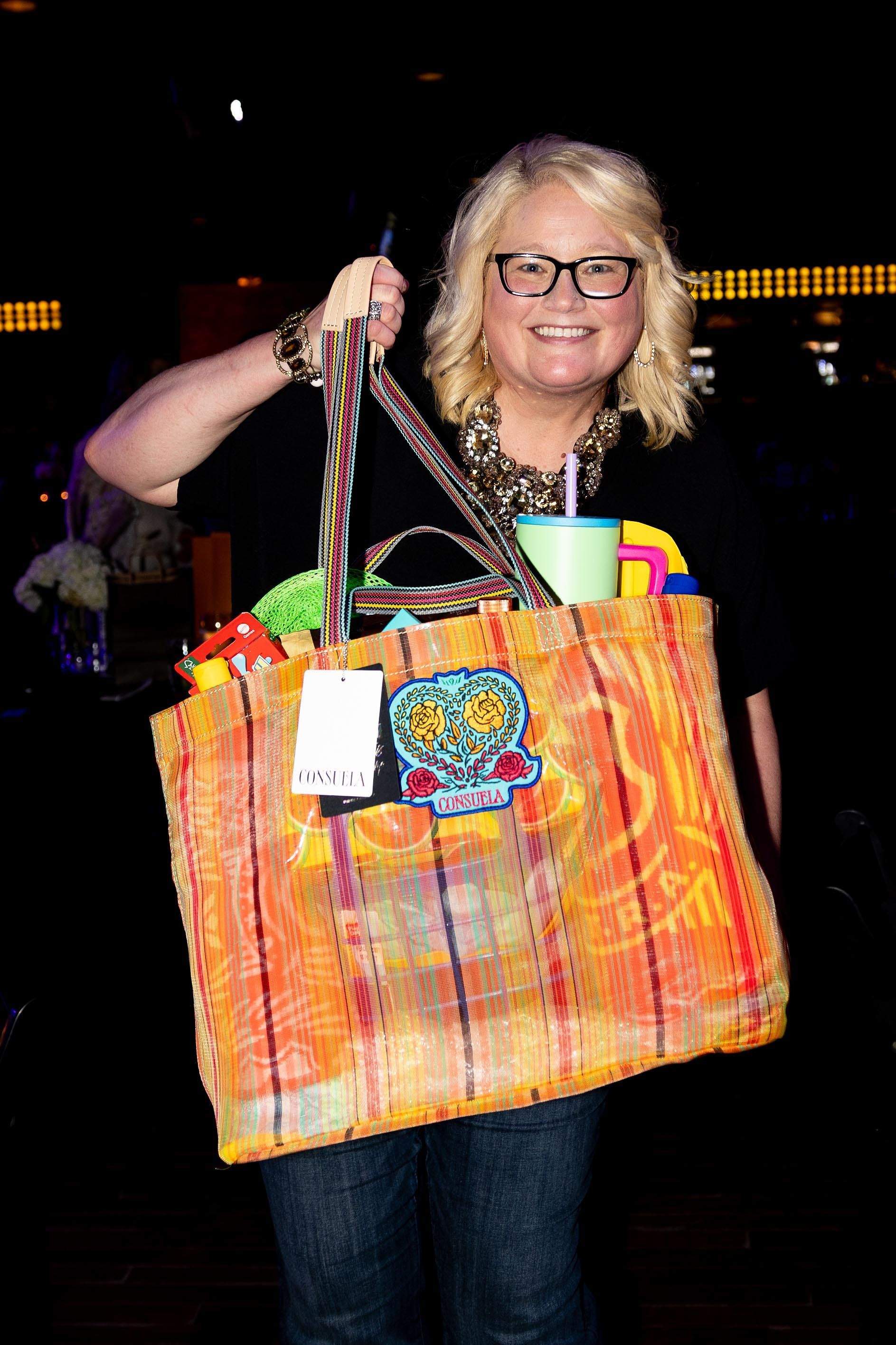 Woman holding a large orange tote bag, smiling. Inside are items like a cup and accessories; nighttime setting.