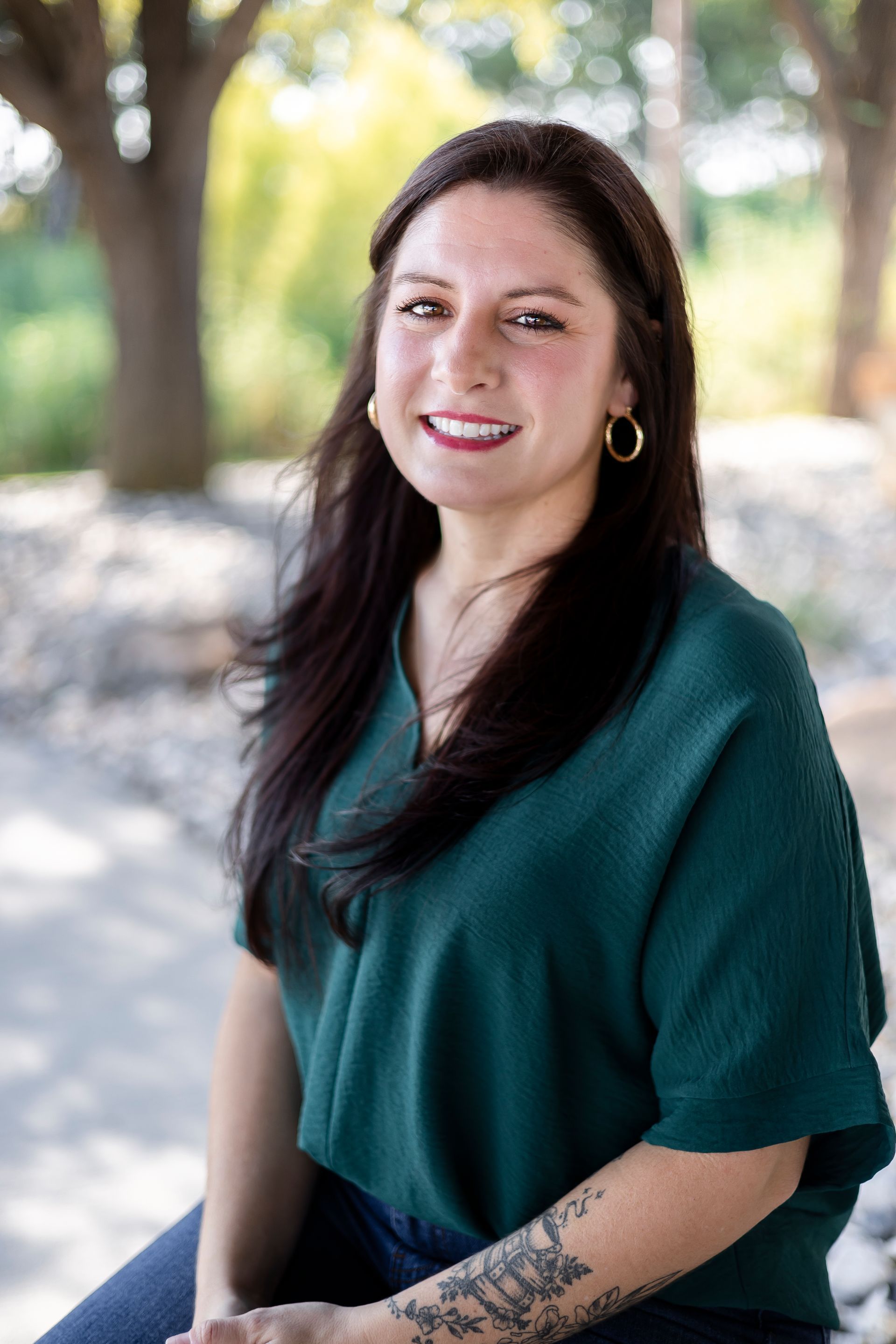 Woman with long dark hair, wearing a teal top and gold hoops, smiling outdoors.