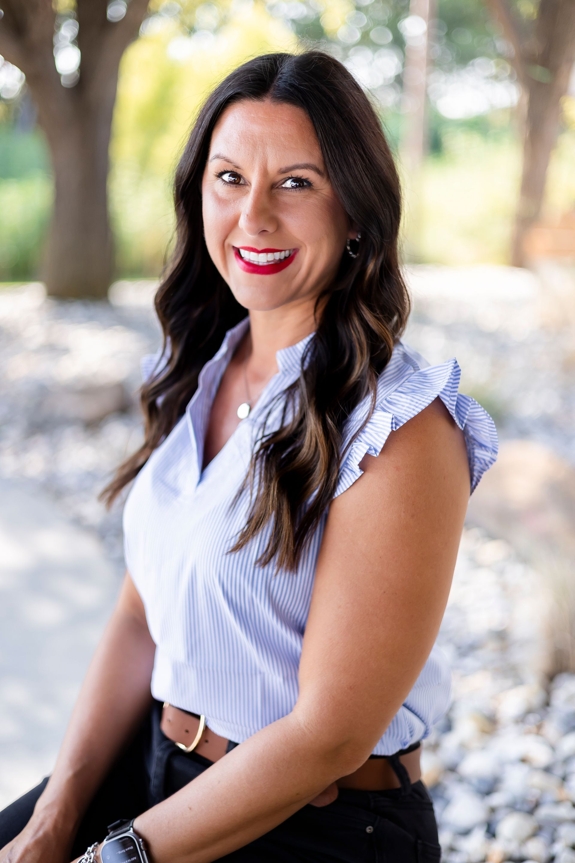 Woman with dark hair and red lipstick smiles, wearing a blue striped top, outdoors.