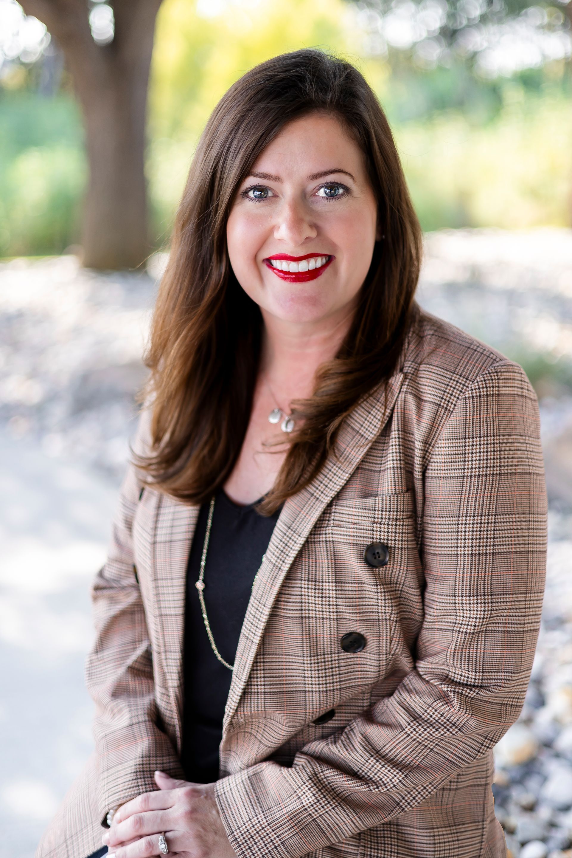 Woman with brown hair smiles, wearing a plaid blazer, black shirt, and necklace, outdoors.