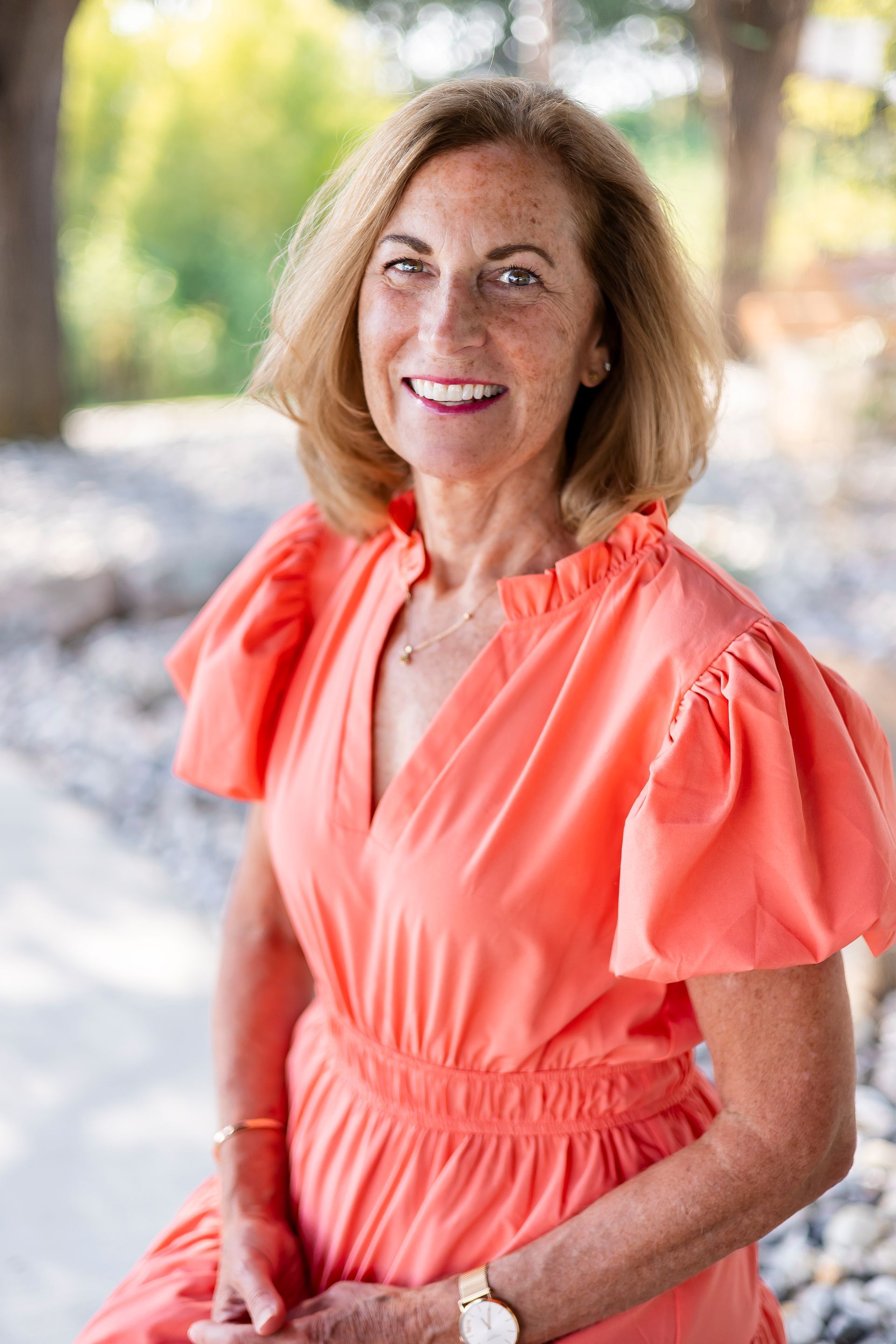 Woman with short blonde hair, smiling in a coral dress outdoors.