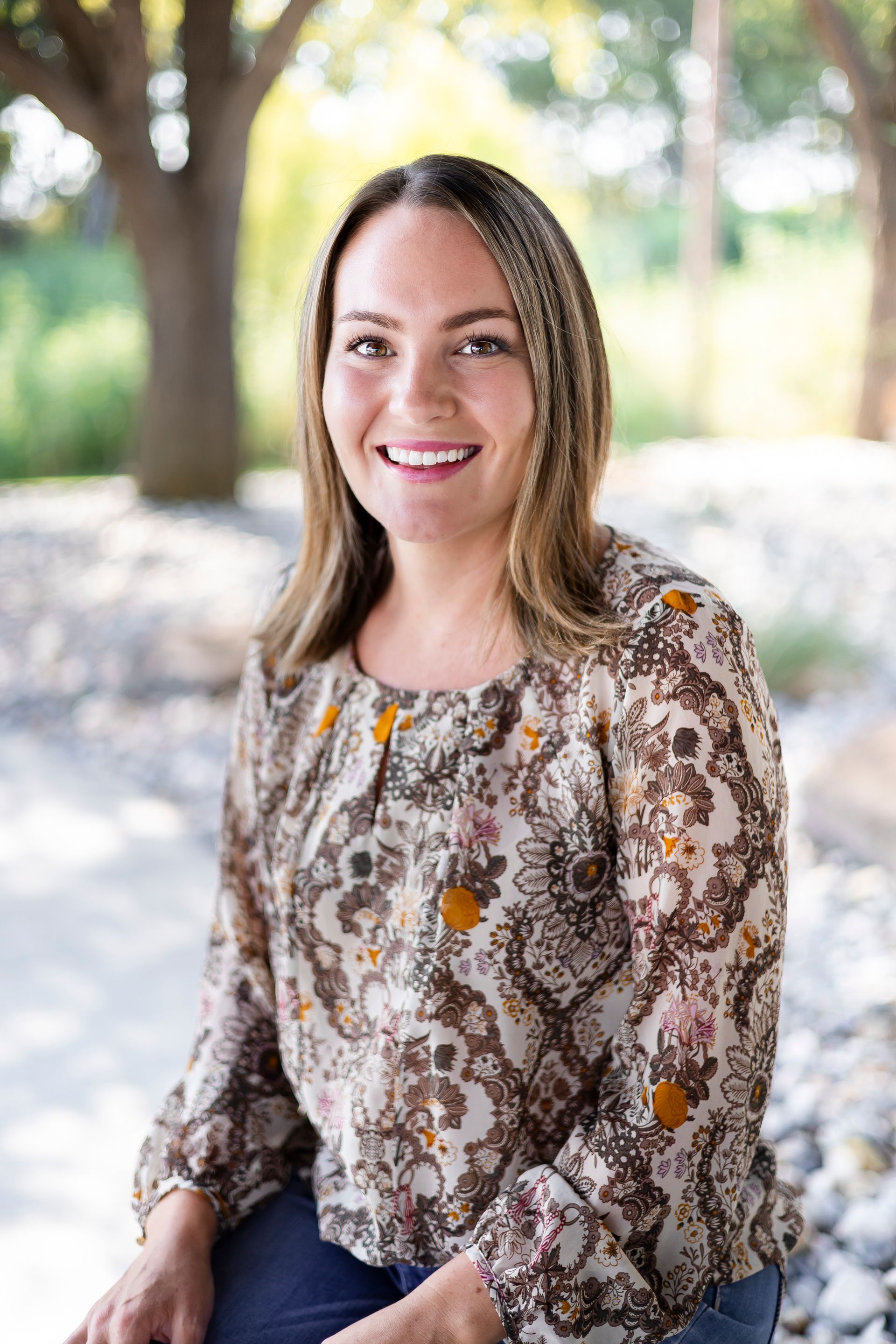 Woman smiling, sitting outdoors, wearing a floral blouse.