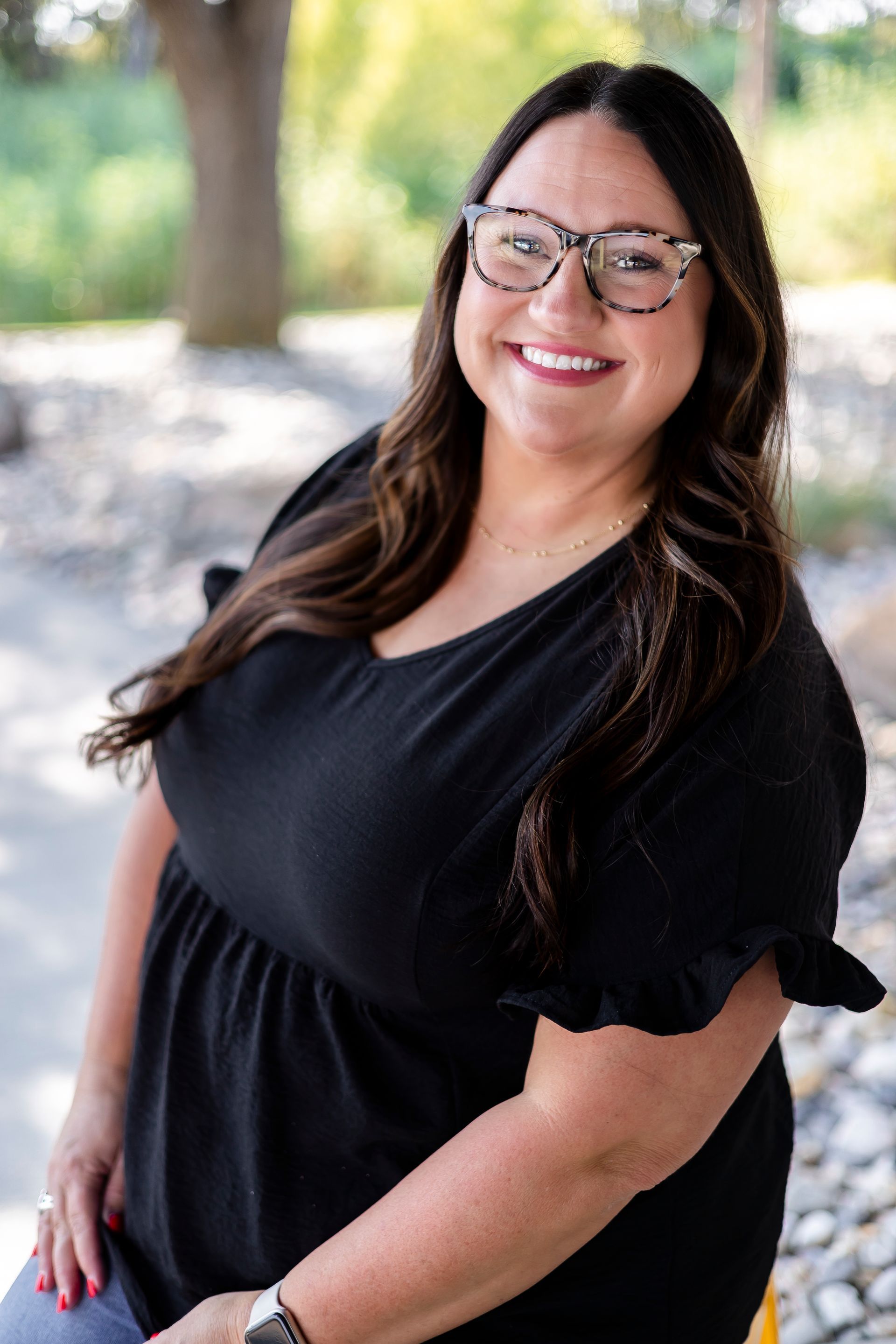 Woman with glasses smiles, wearing a black top outdoors.