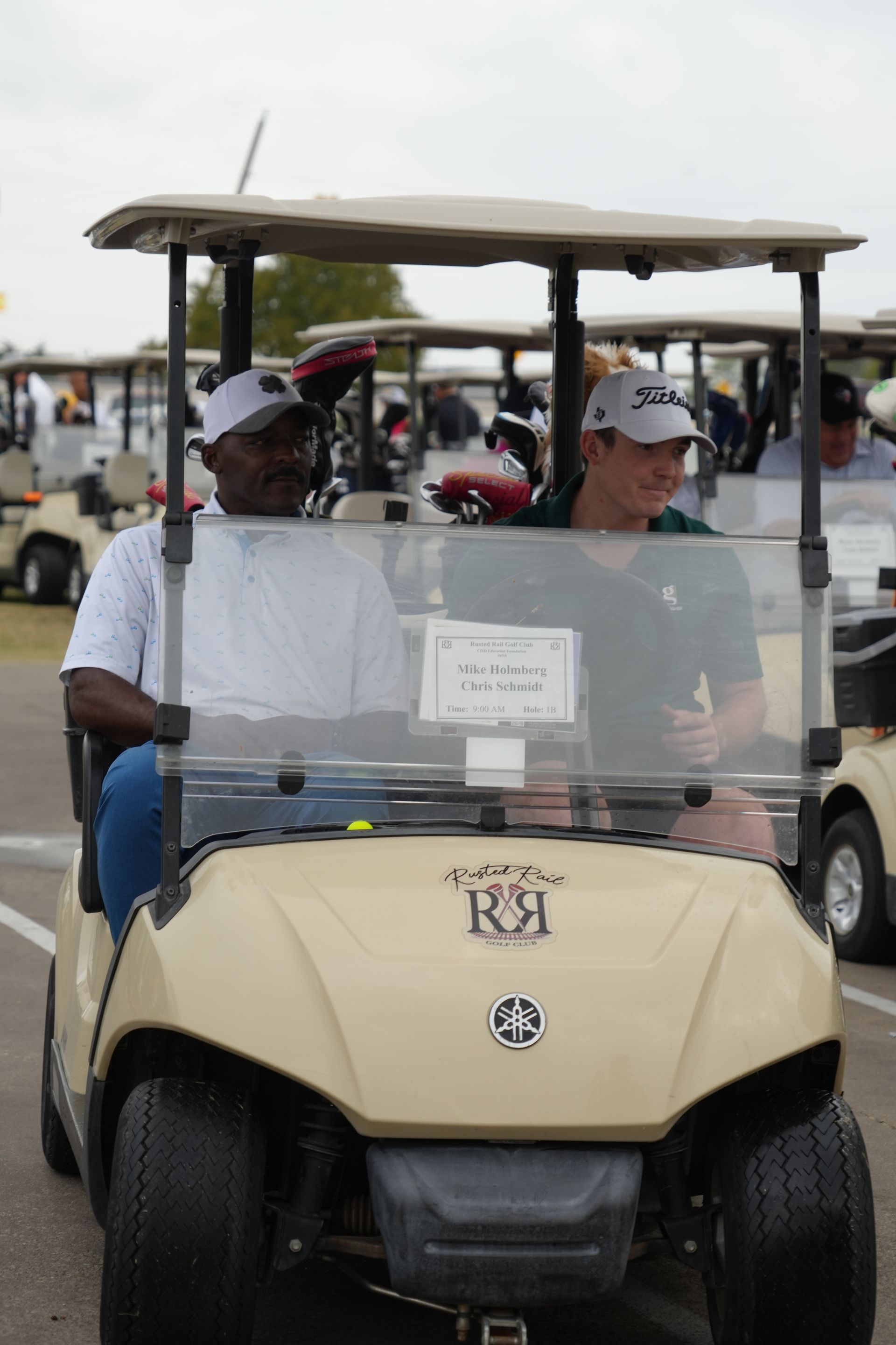 Two men in a tan golf cart; one black, one white, smile; other golf carts in background.