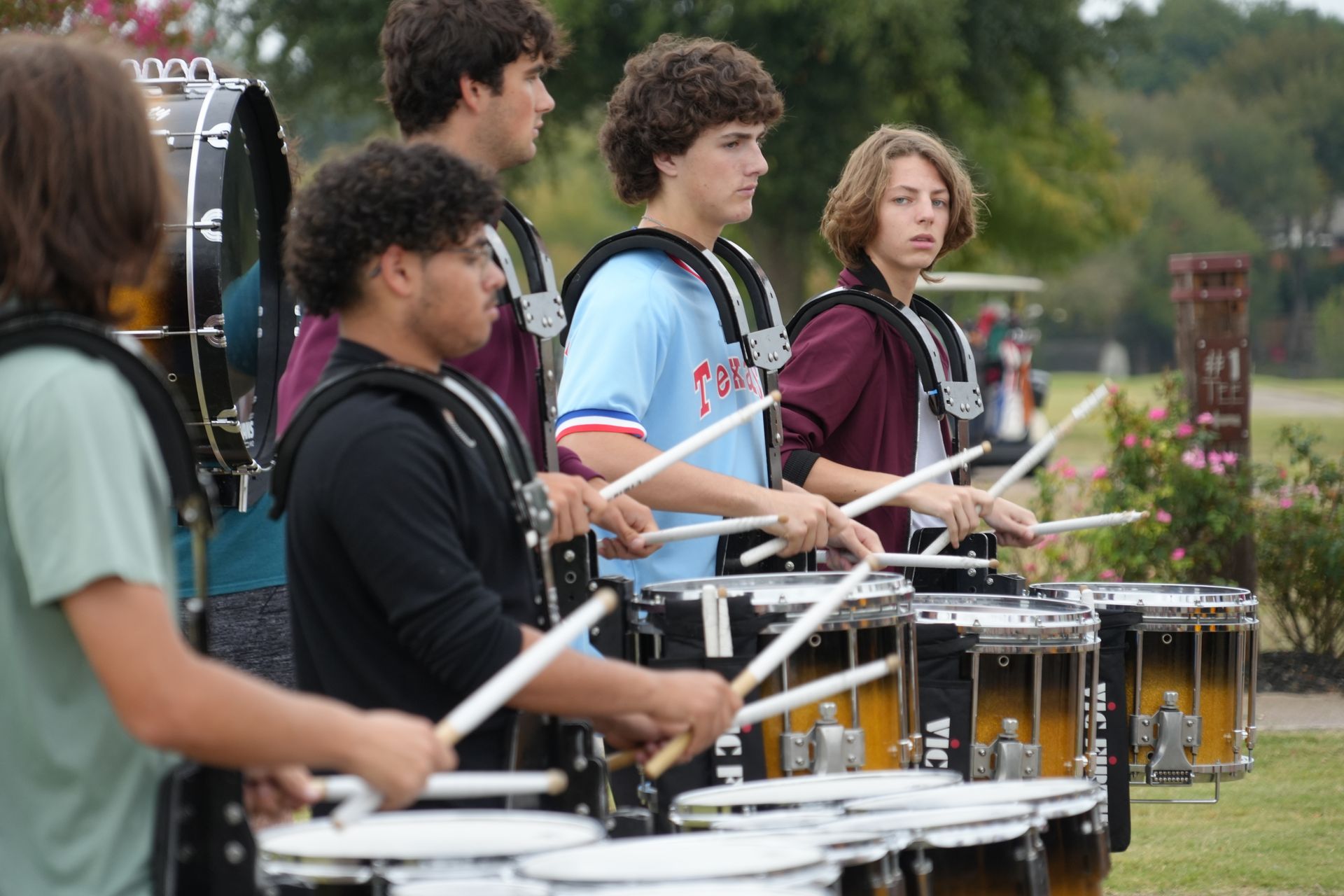 Marching band drumline performing outdoors with several drummers, wearing various outfits.