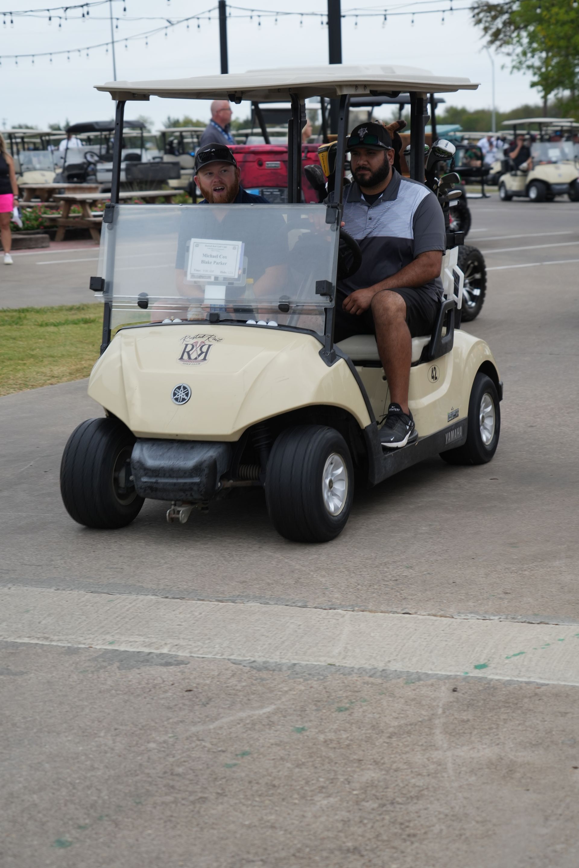 Two men in a beige golf cart driving on a paved area.