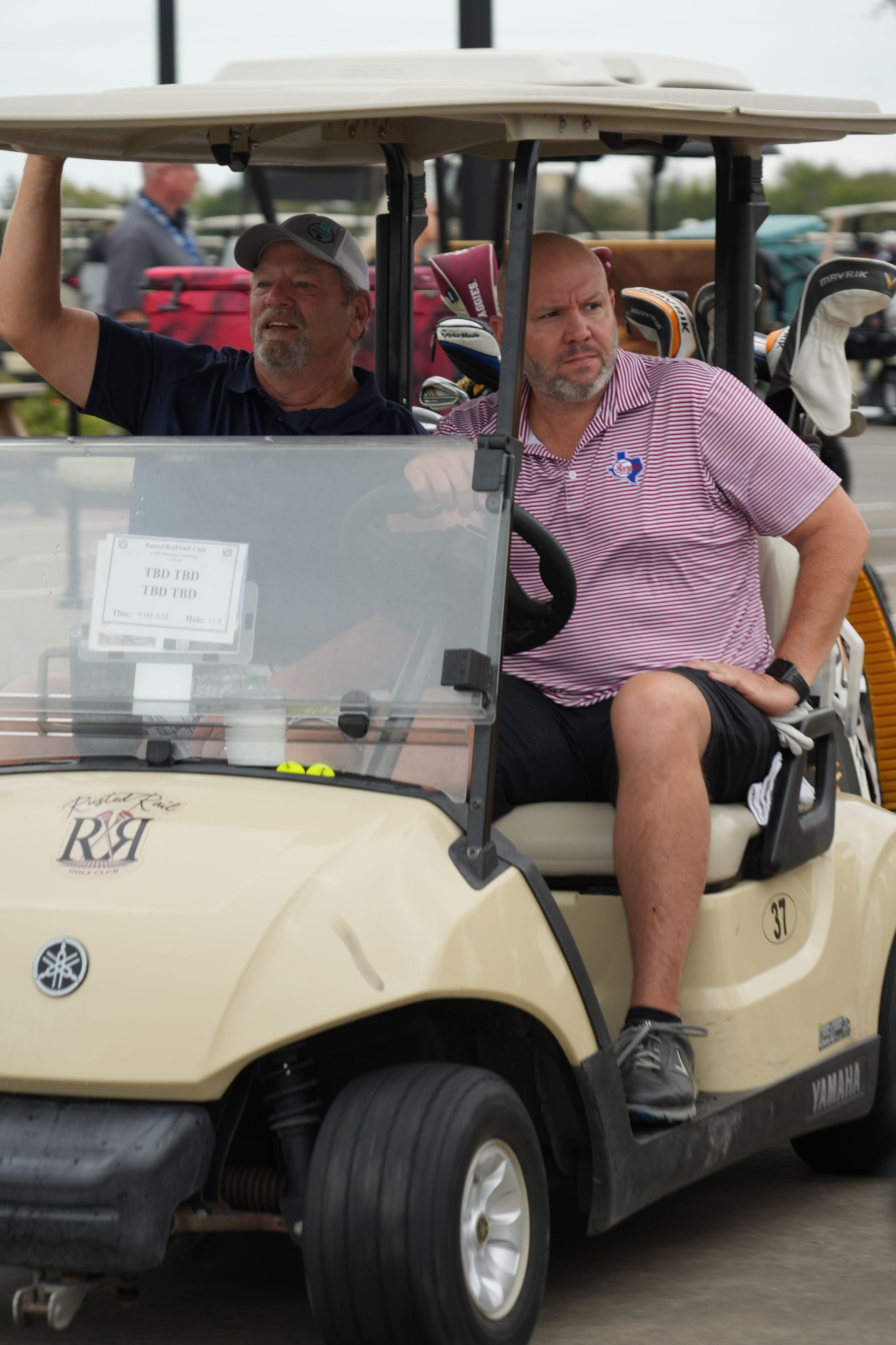 Two men in a beige golf cart; one waves, the other looks ahead. Outdoors, likely on a course.