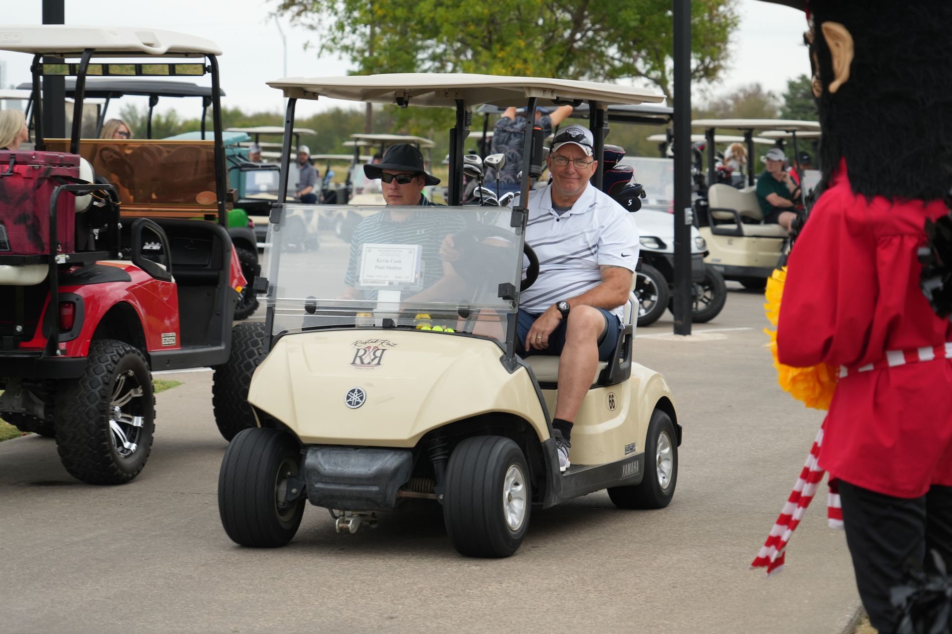 Men in golf cart, smiling. Other golf carts and a mascot in the background. Outdoors.