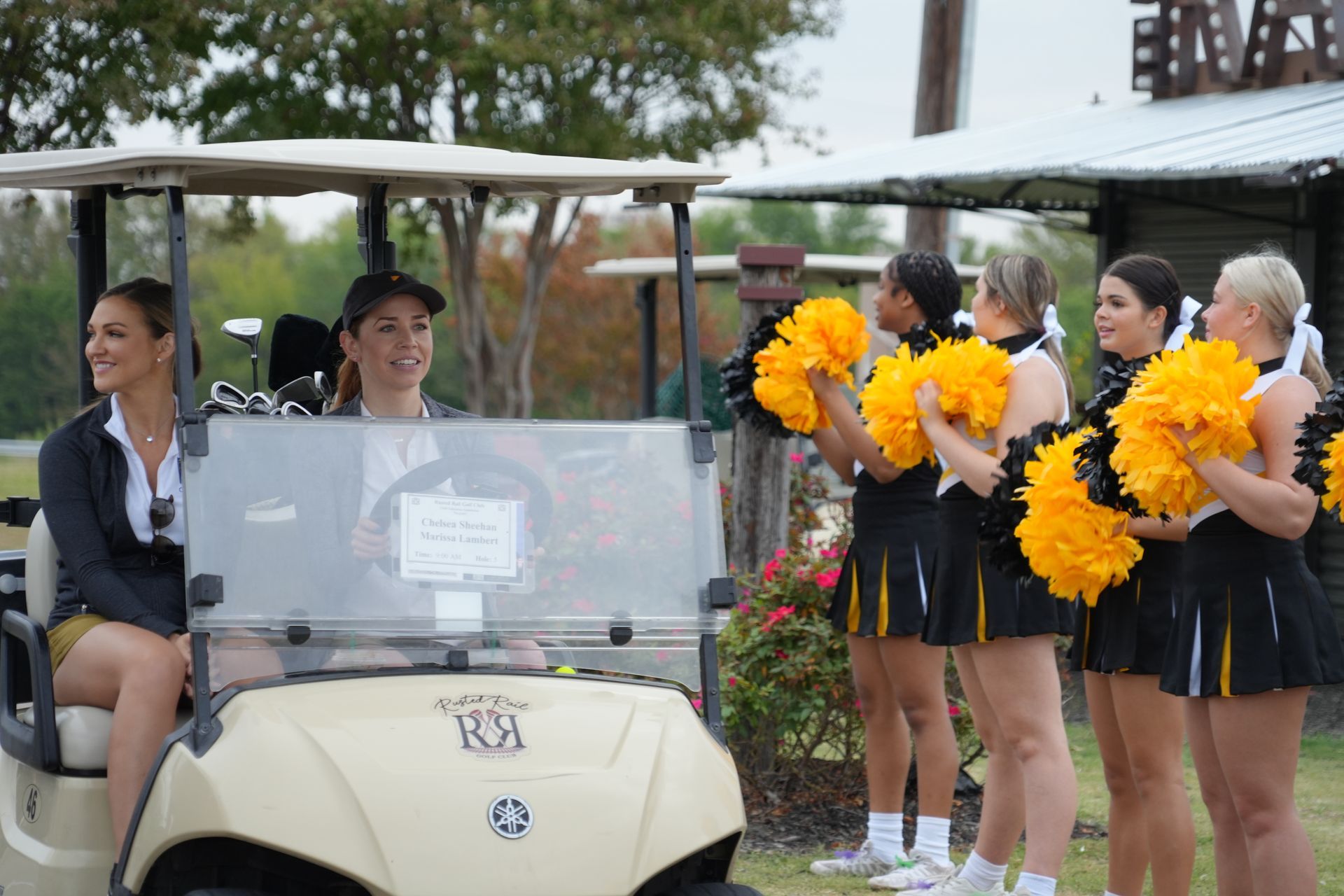 Two women in a golf cart greeted by cheerleaders with yellow pom-poms.