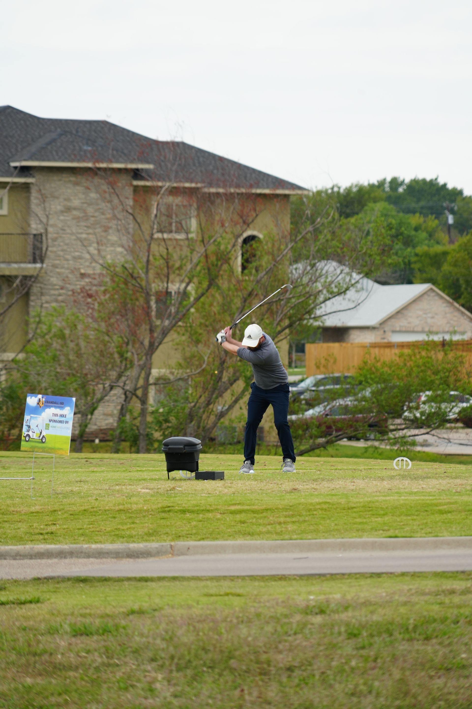 Golfer in mid-swing on a green, wearing a cap and dark clothes, with houses and cloudy sky in the background.