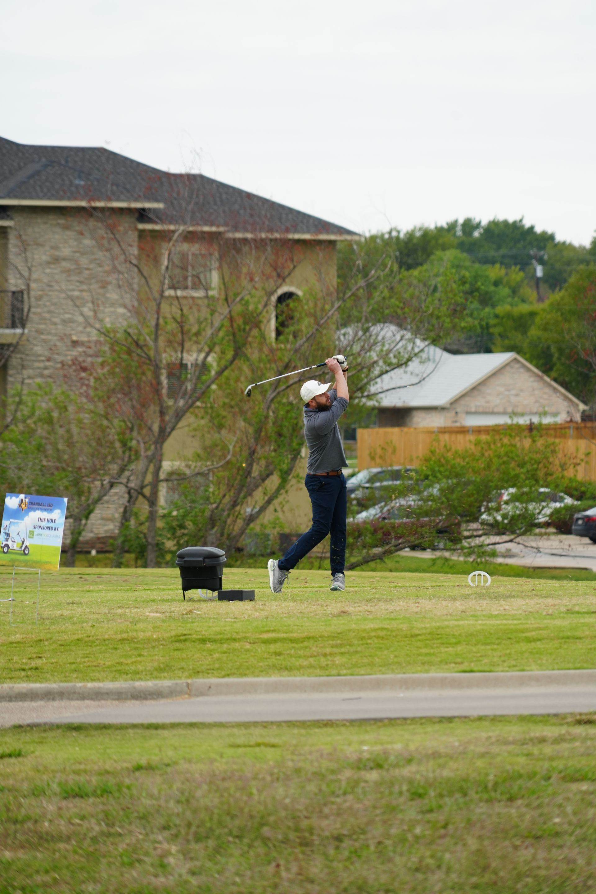 Golfer in dark pants and white hat swings club on a green, with a residential building in the background.