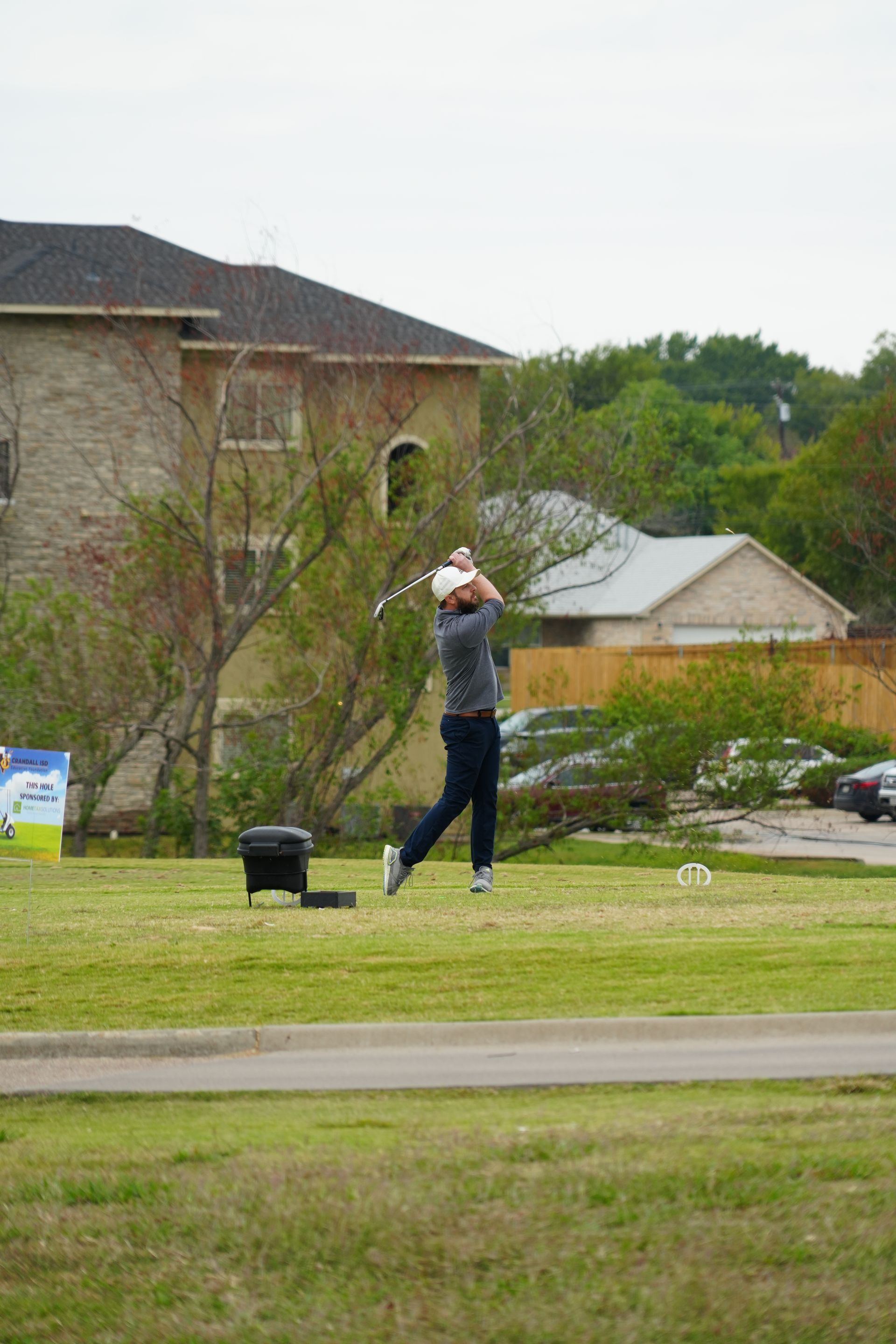 Golfer swinging on a green, in front of a building and trees on a cloudy day.