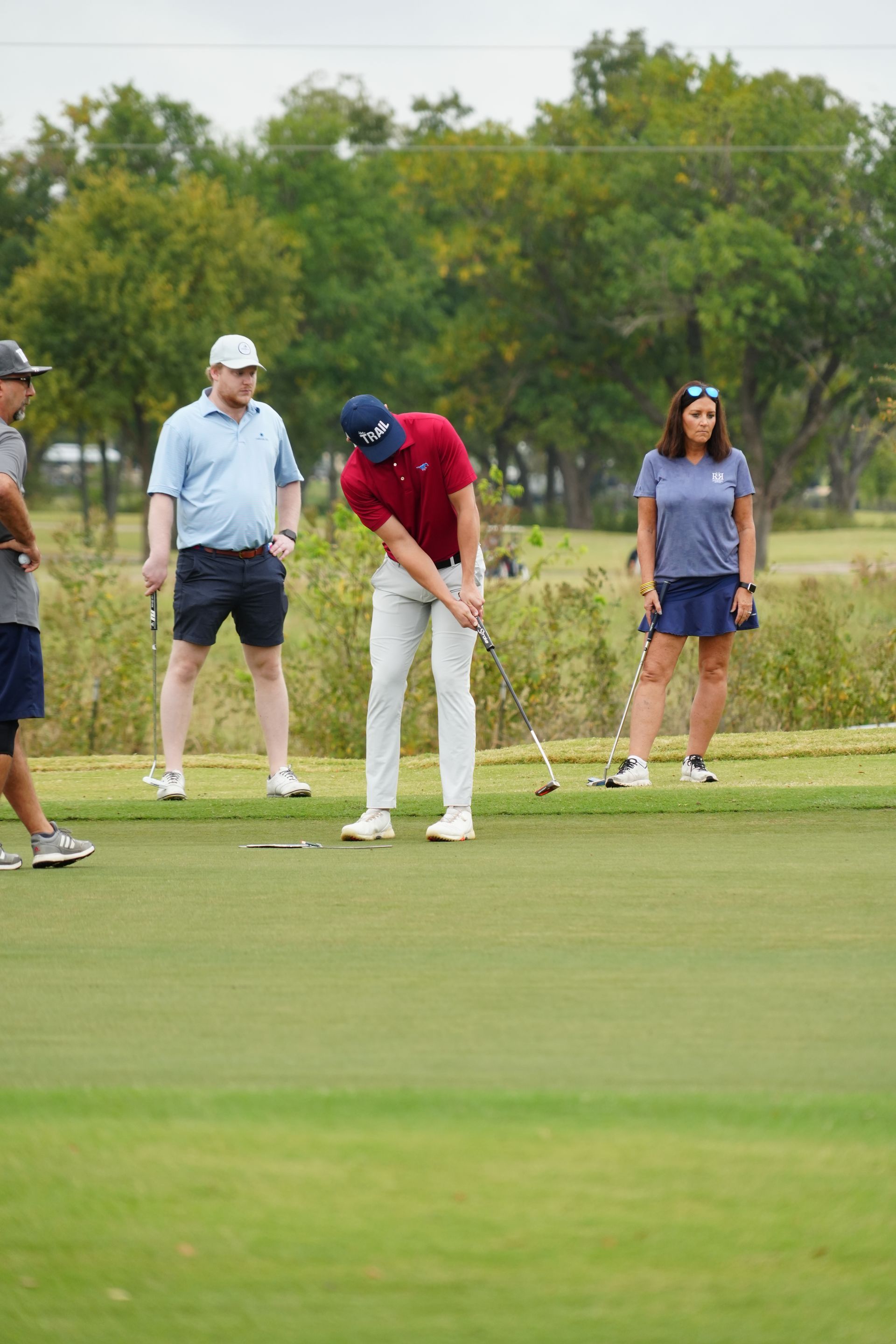 A golfer in red shirt and white pants putts on a green, observed by others at a golf course.