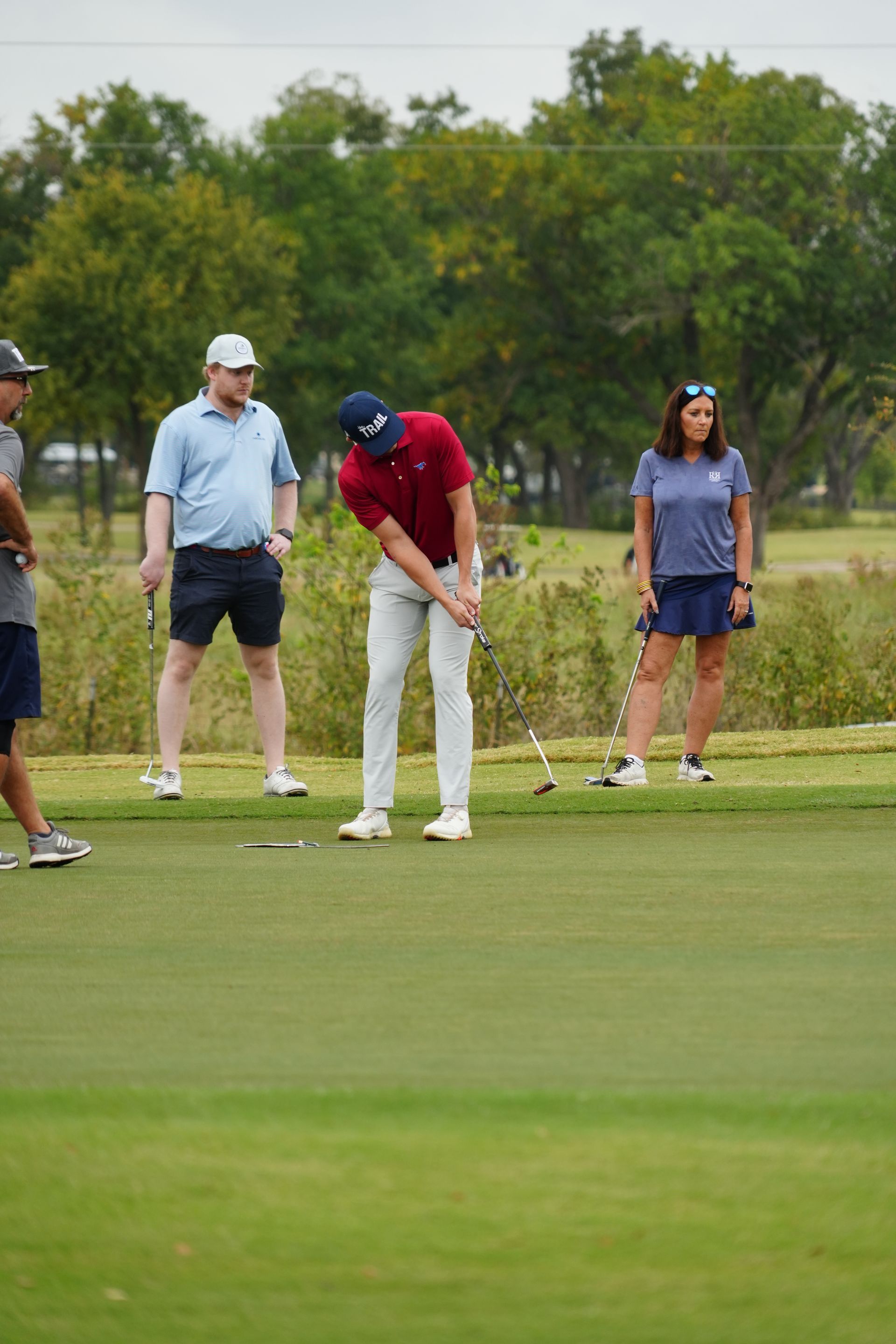 Man in red shirt putting golf ball, watched by three others on green, trees in background.