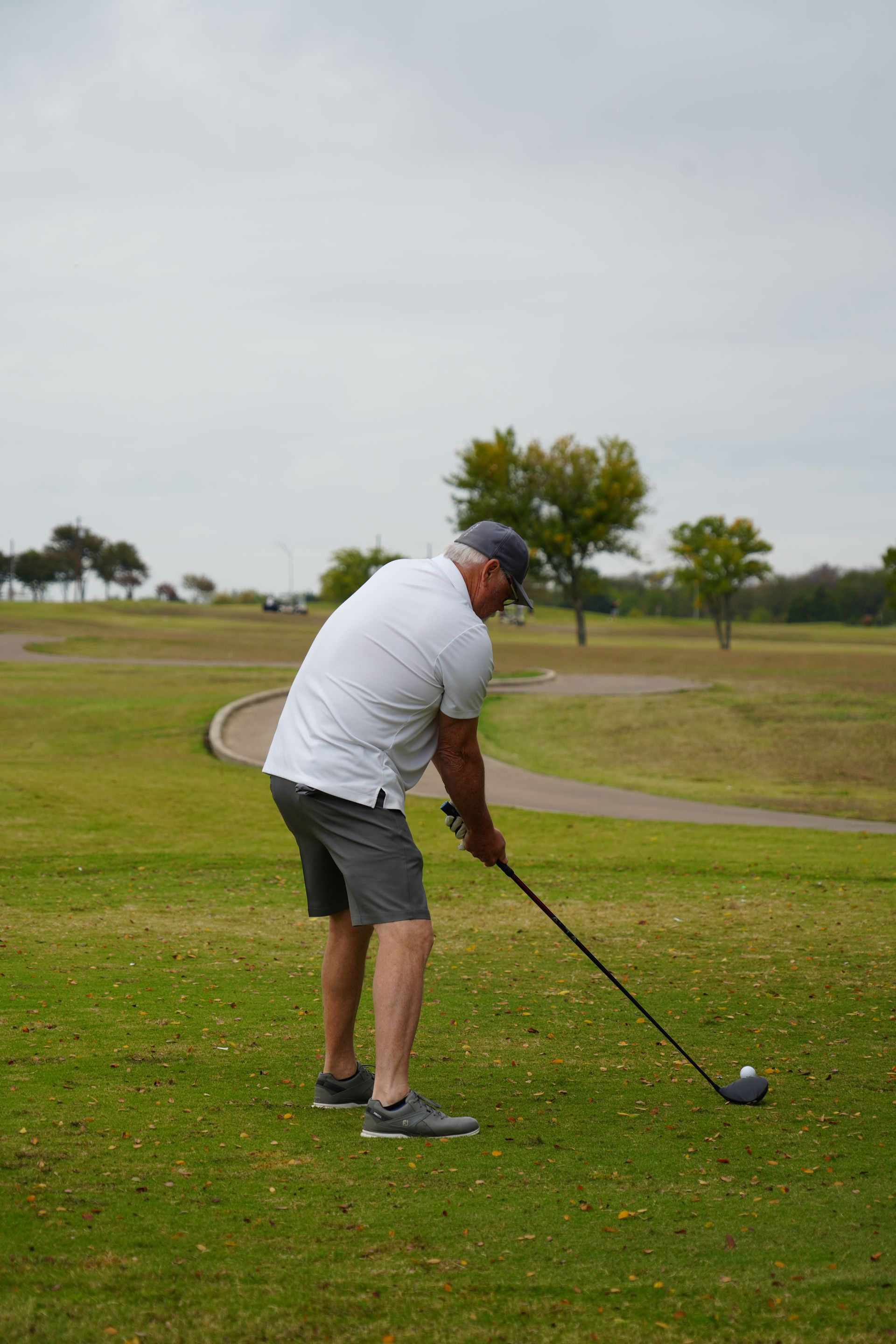 Man in white shirt and shorts tees off on a golf course, under an overcast sky.