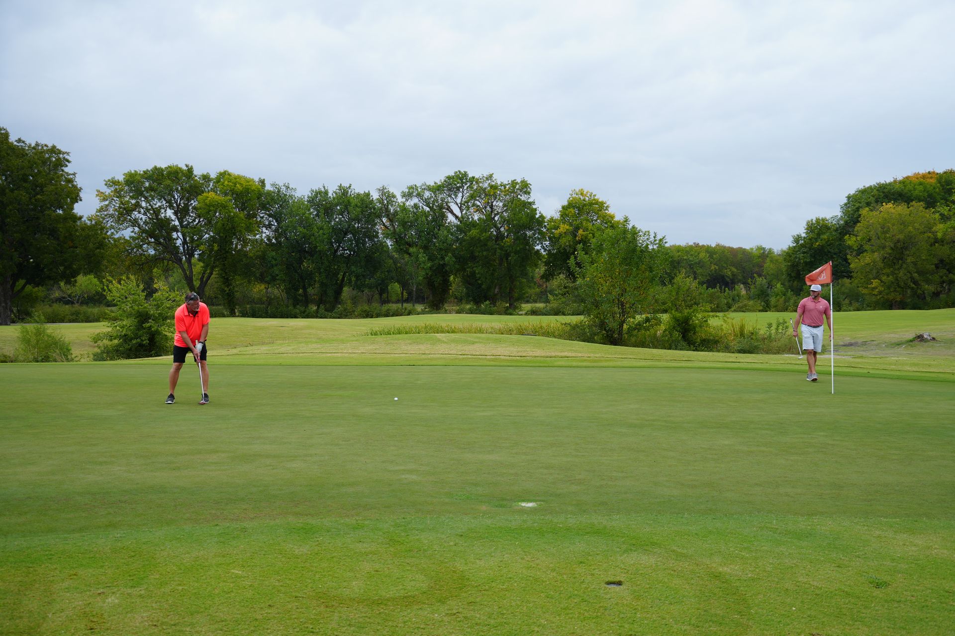 Two golfers on a green, one putting, the other holding the flag on a cloudy day.