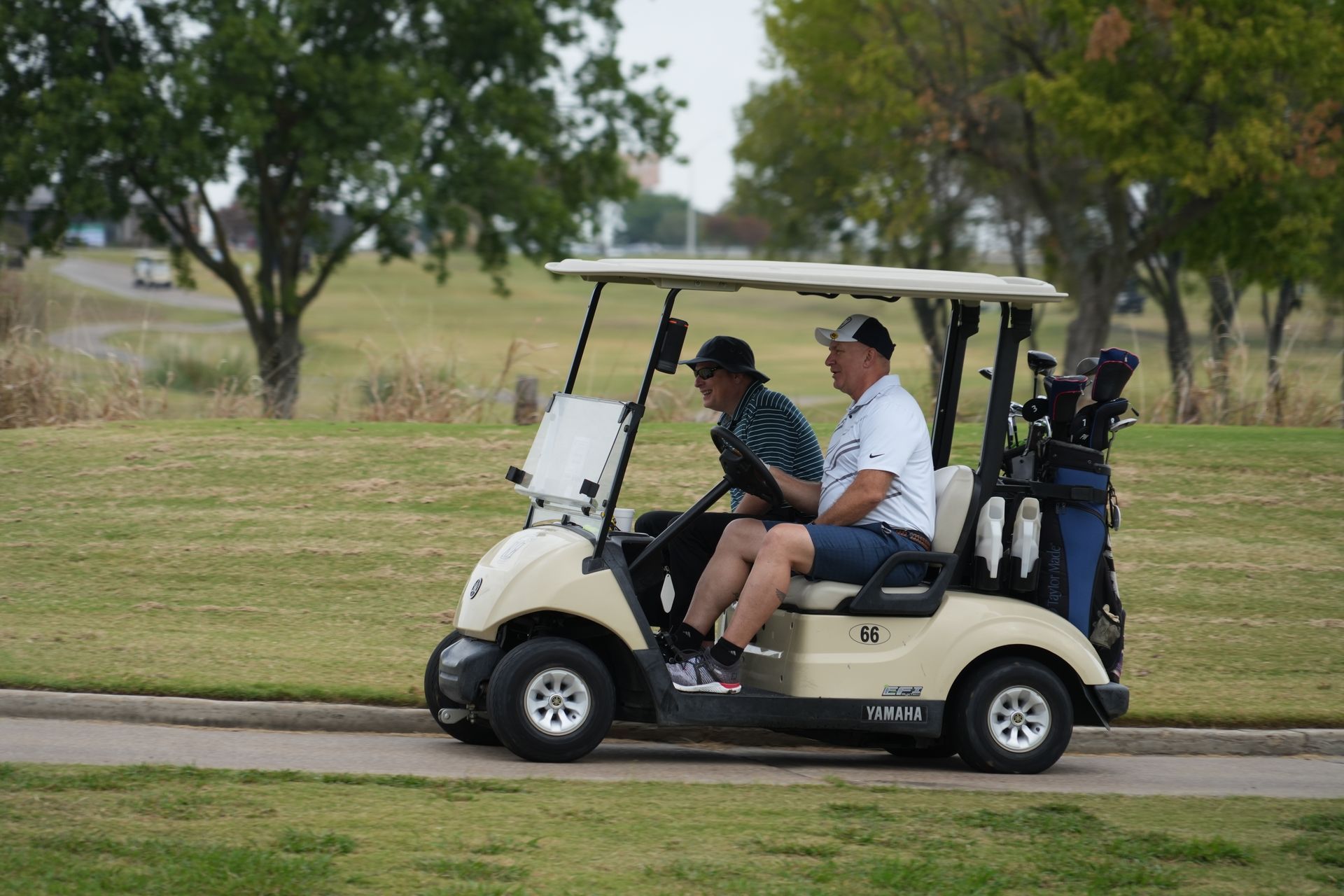 Two men in a beige golf cart on a course, golf bags in back, one driving.