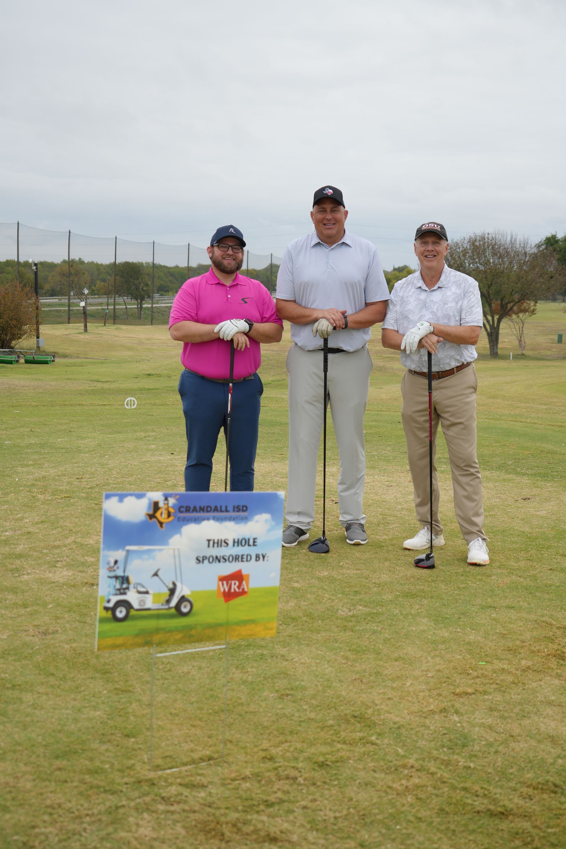 Three men on a golf course, smiling with golf clubs. Sign in front. Overcast day.