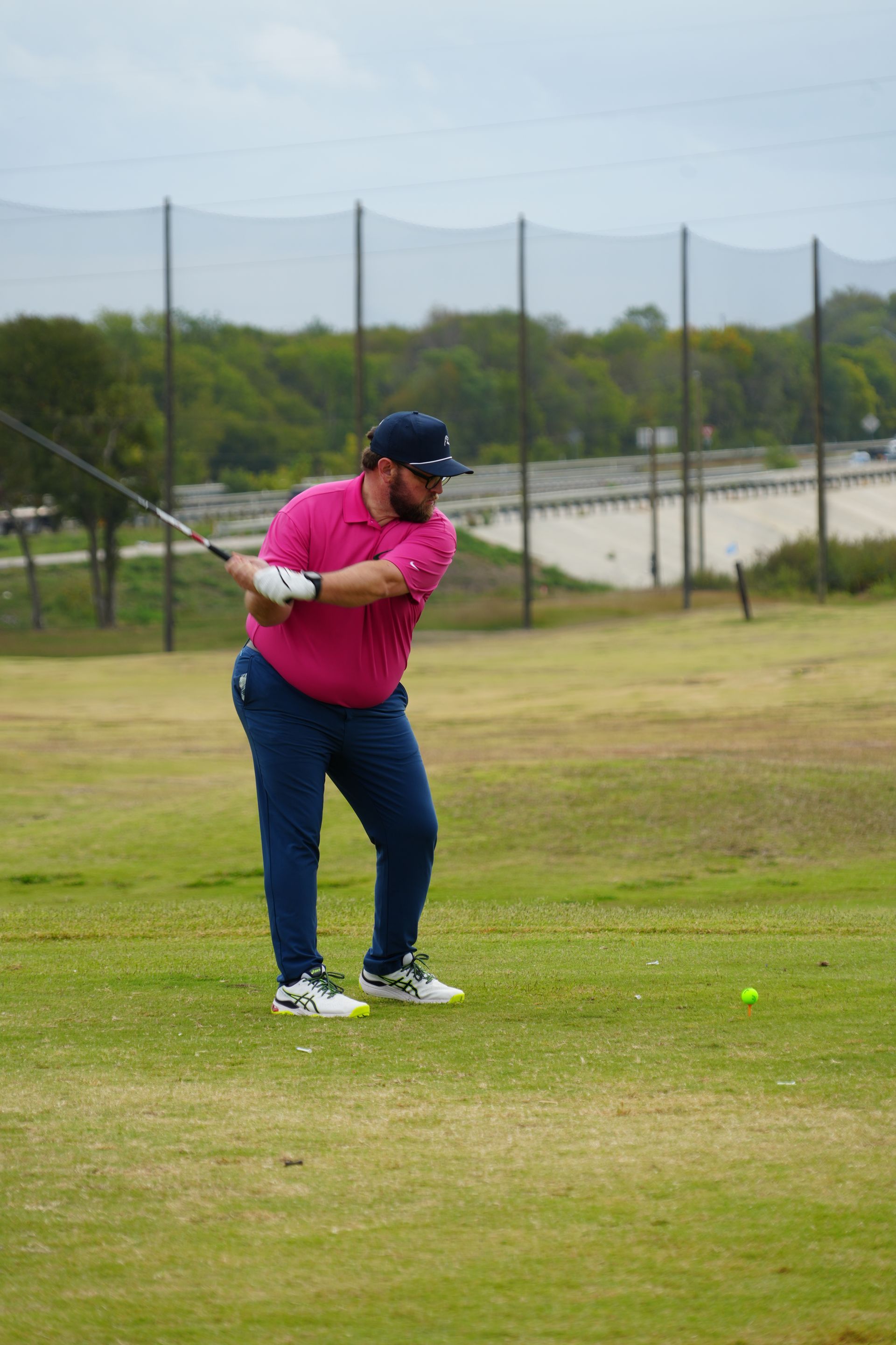 Man in pink shirt swings a golf club on a driving range; green grass, blue pants, and netting in the background.