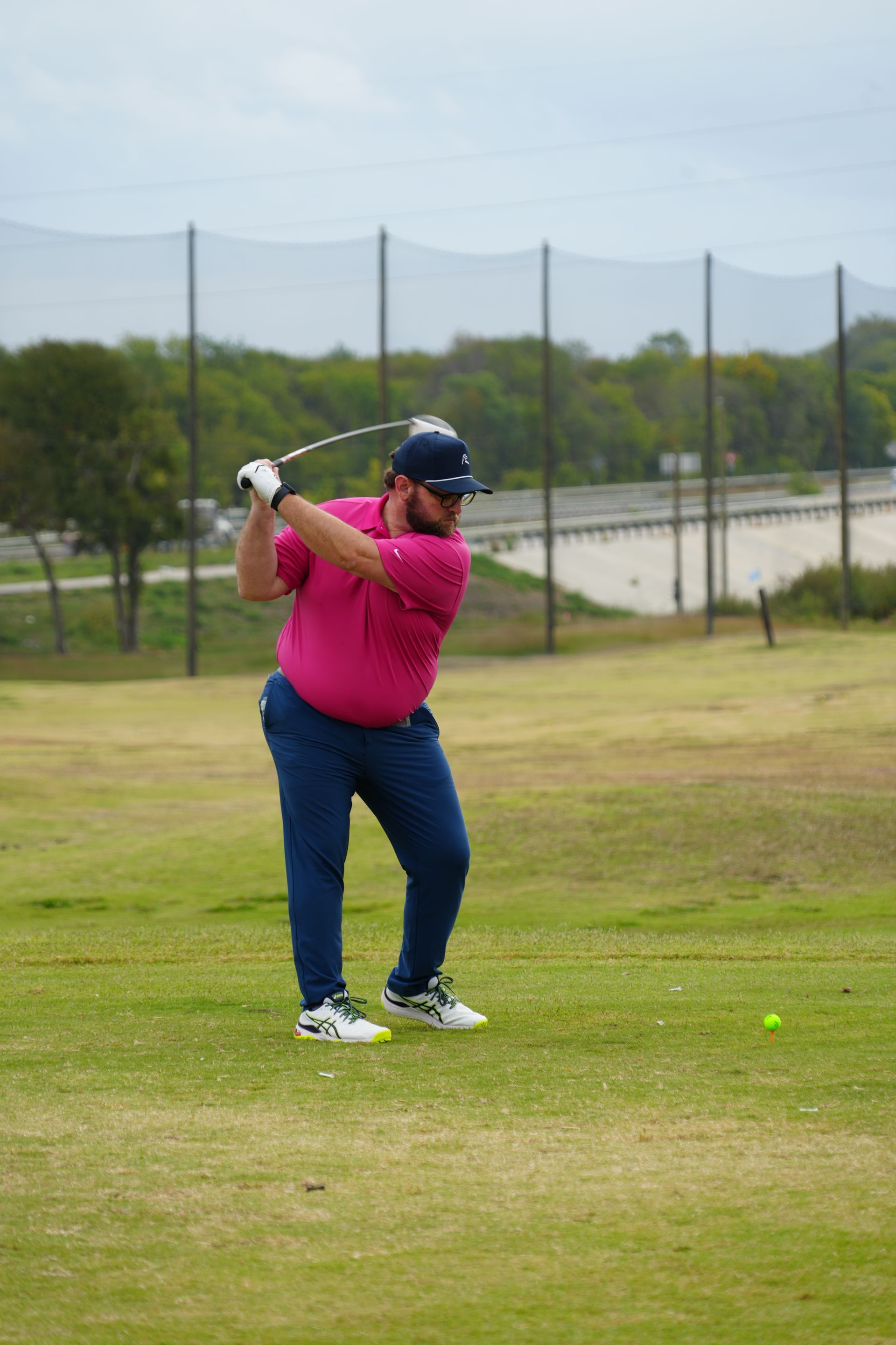 Man in pink shirt and blue pants swings golf club on a driving range.