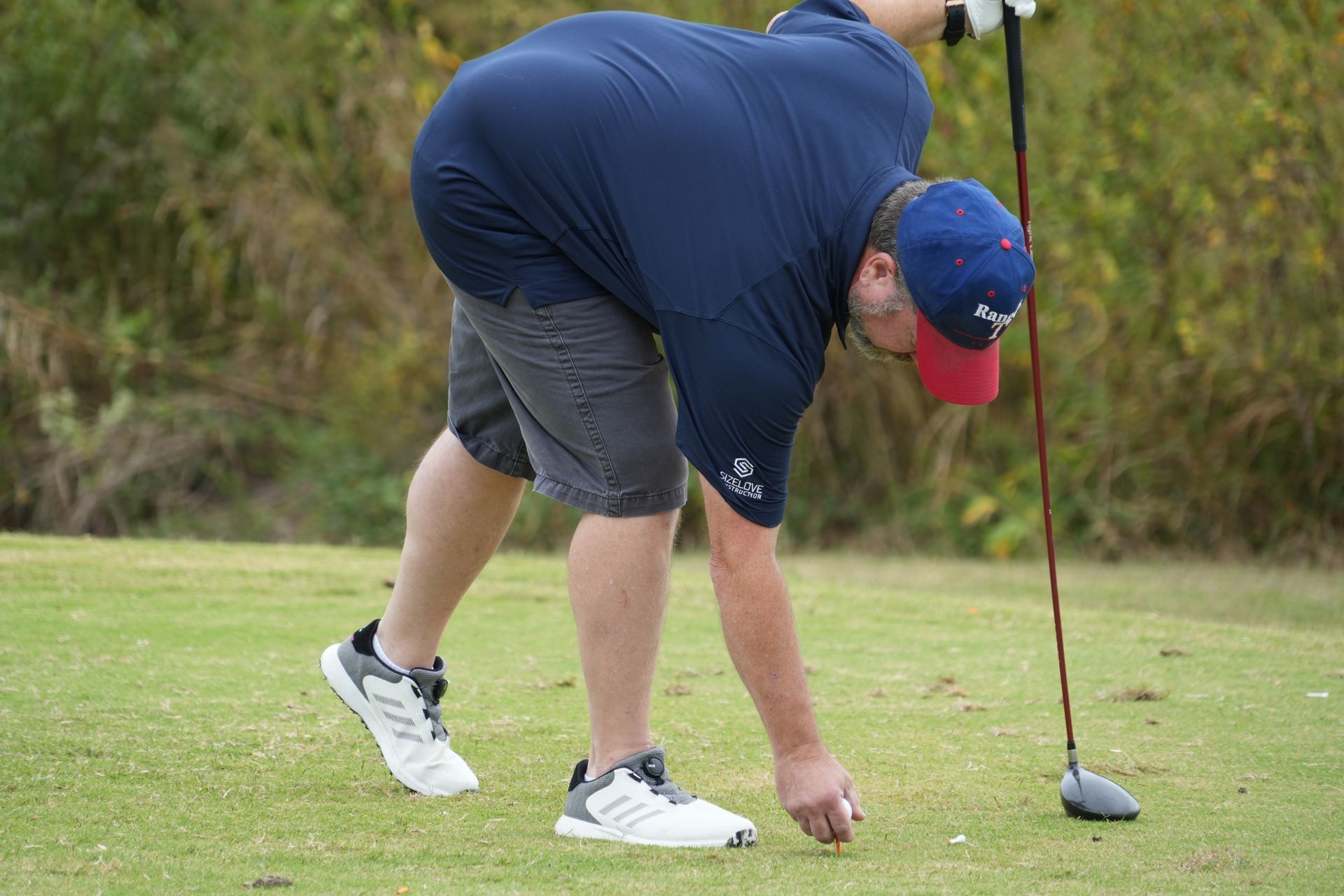 Man in navy shirt and shorts on a golf course, picking up a golf ball, near a tee.