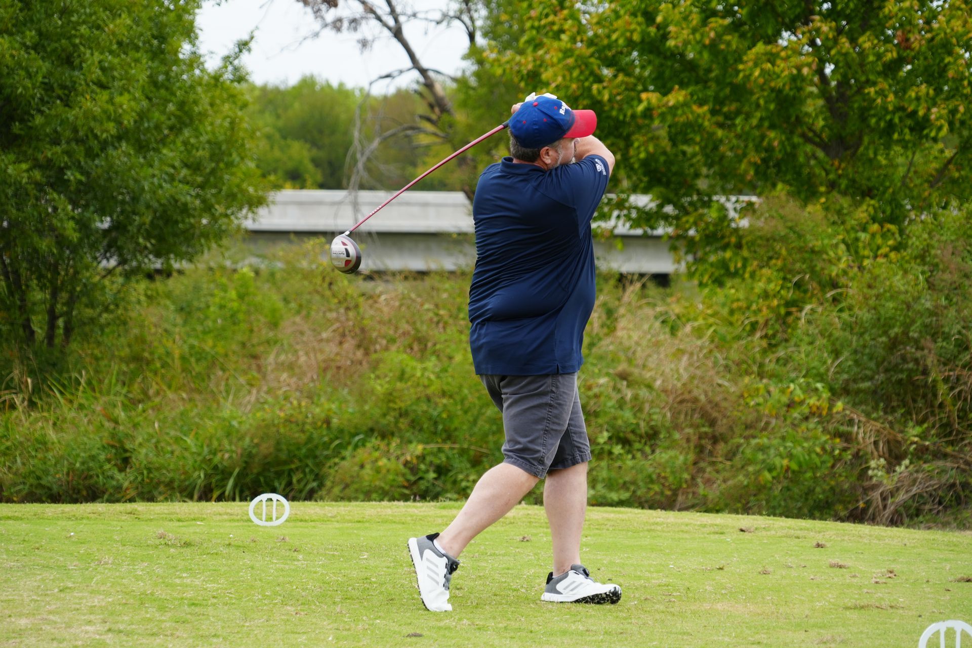 Person in blue shirt and hat swings a golf club on a green course.