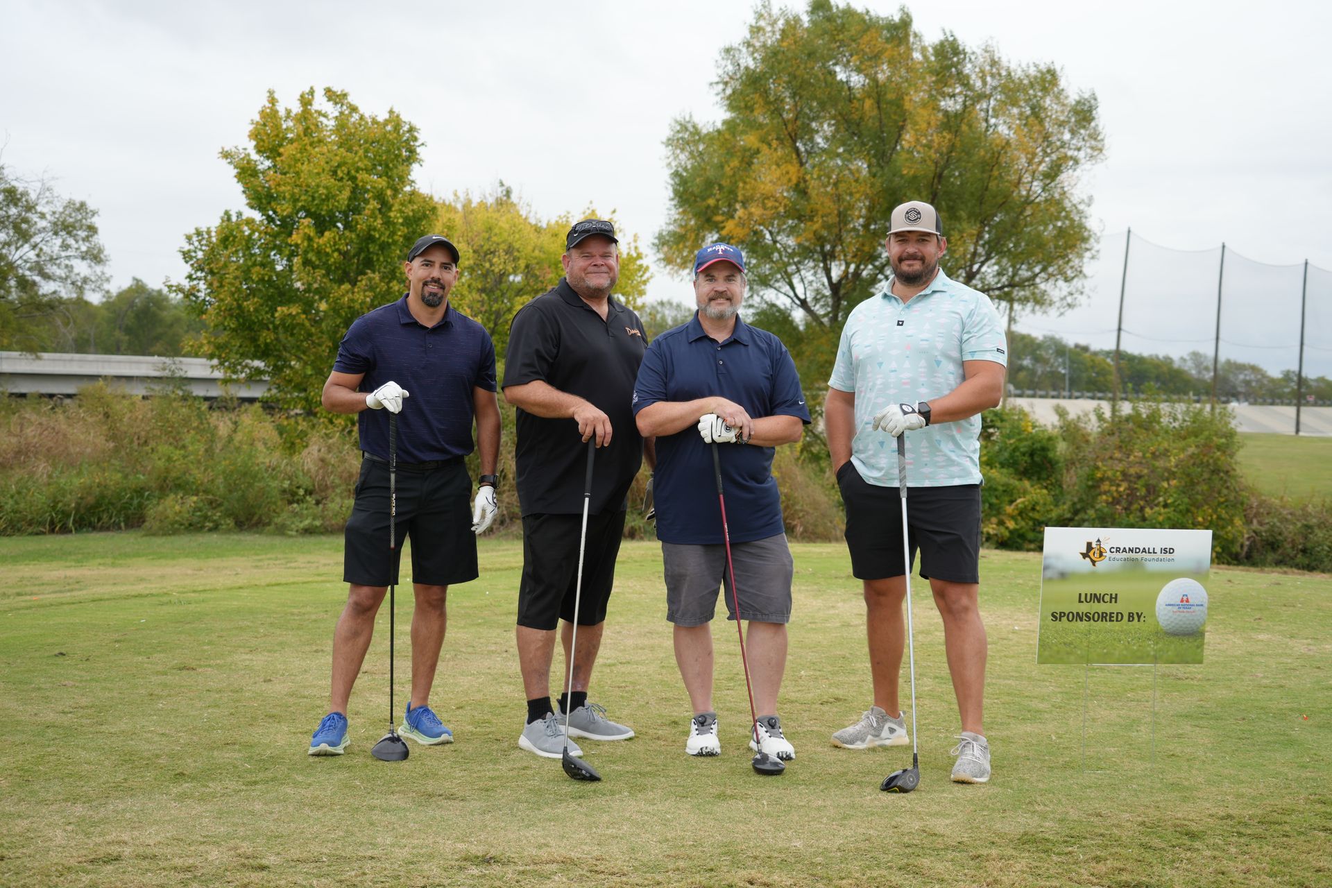 Four men on a golf course, holding clubs. They are wearing shorts and smiling. There is a sign in the background.