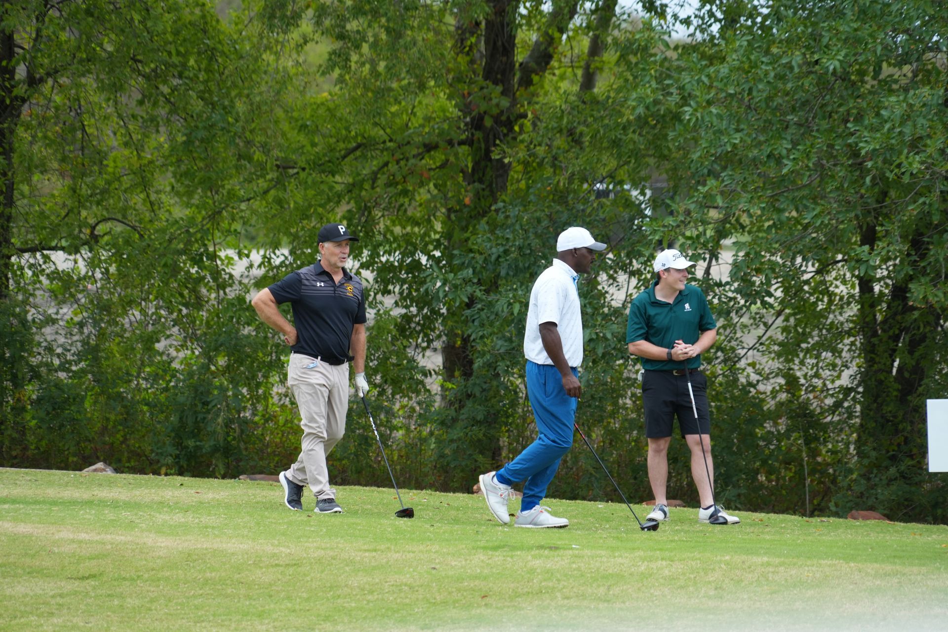 Three golfers on a green: one in black shirt, tan pants; one in white shirt, blue pants; one in green shirt, black shorts.