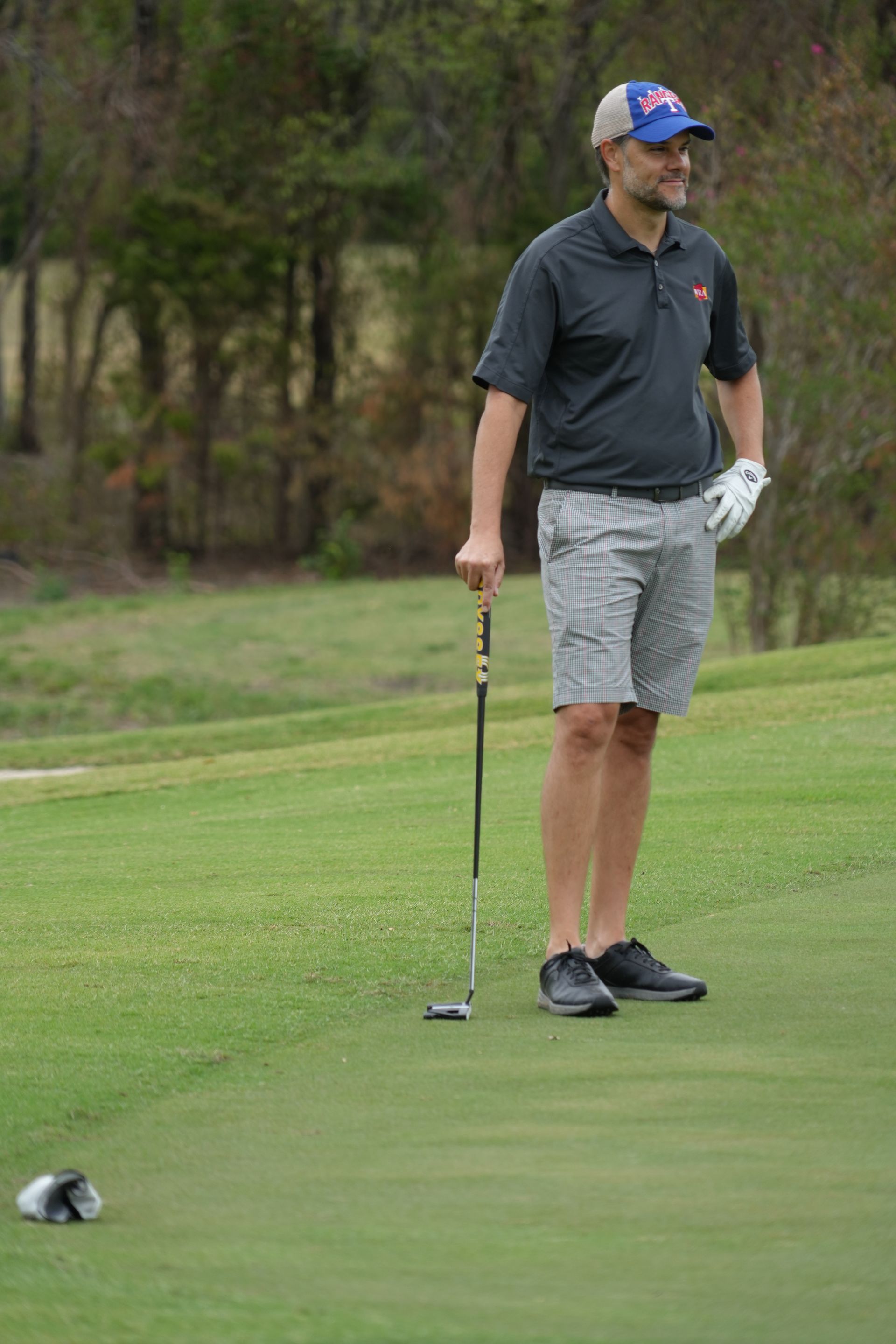 Man in a cap and shorts on a golf course, holding a putter, looking off to the side.