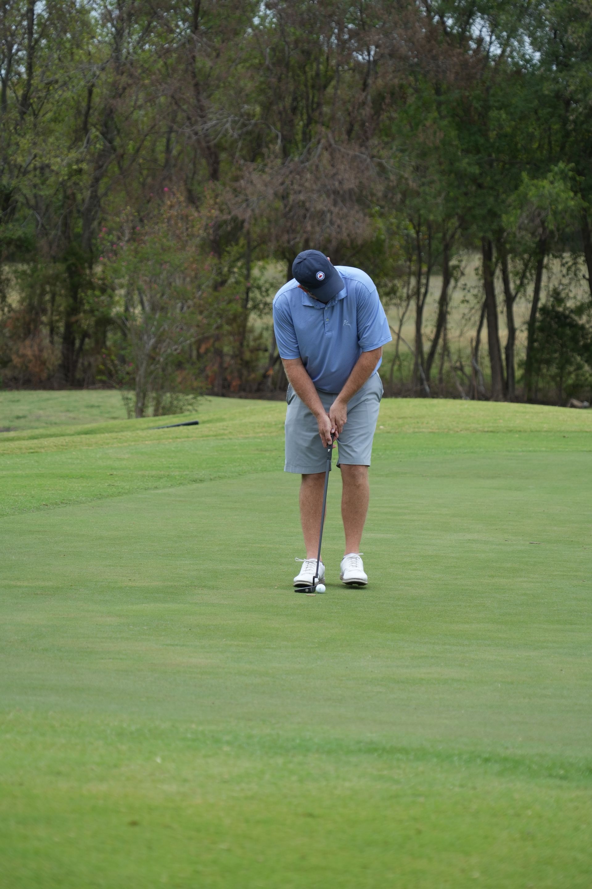 Man in blue shirt, cap, and shorts putting golf ball on green.